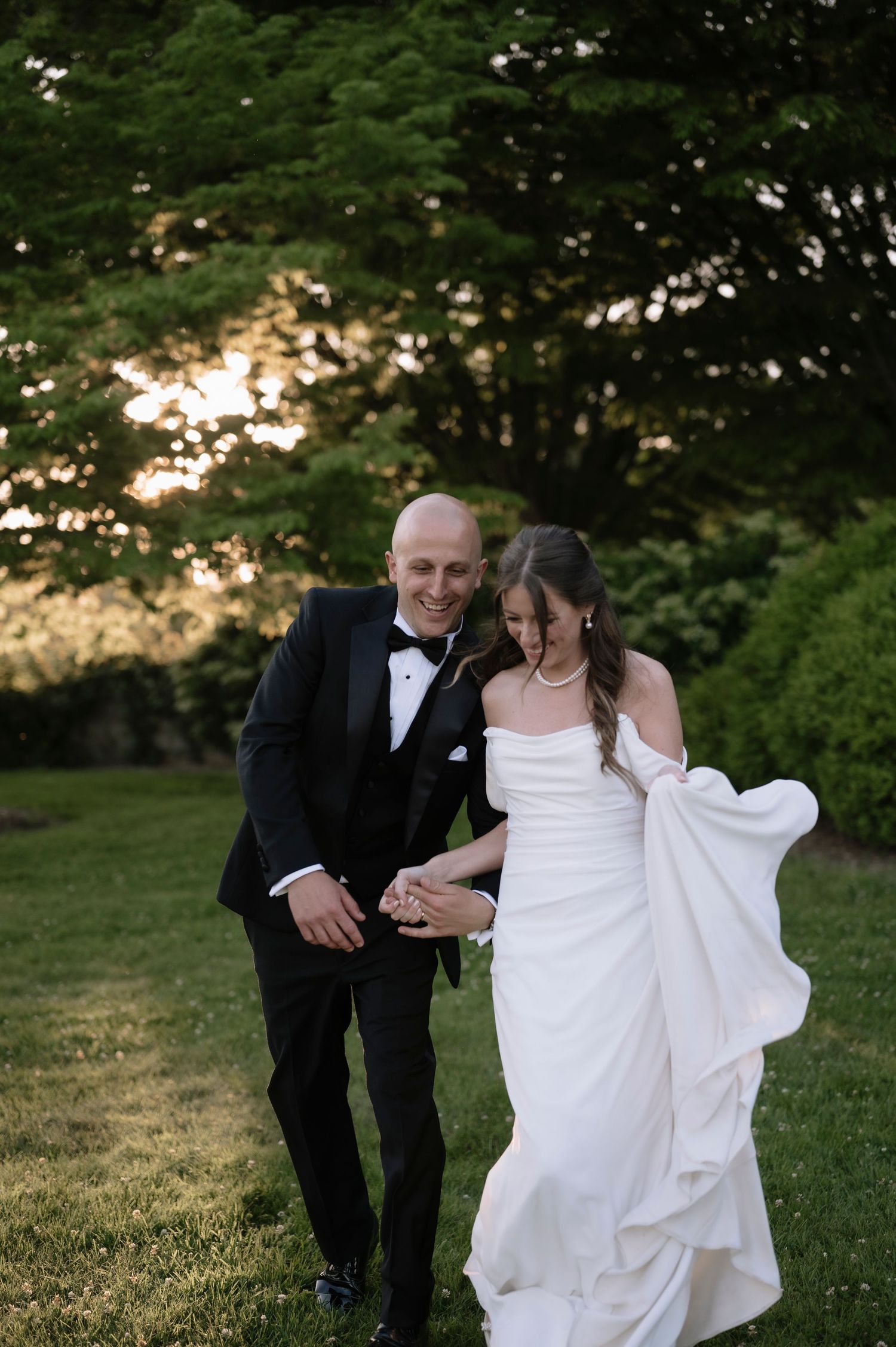 A happy couple walks together on a grassy lawn at sunset during their wedding celebration.