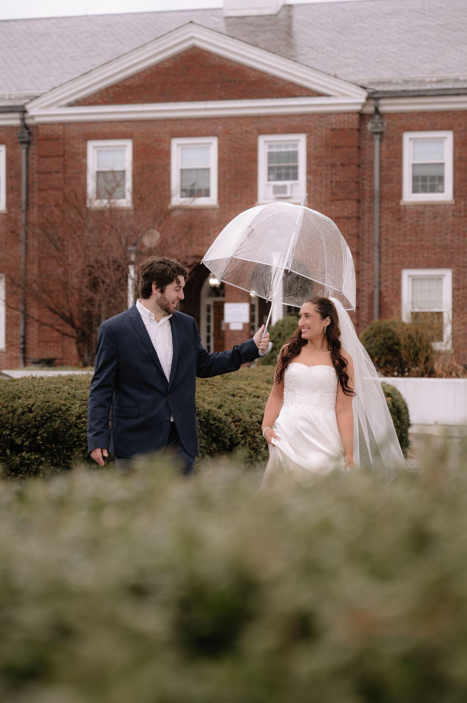 Rainy wedding day portrait with clear umbrella in front of brick building.