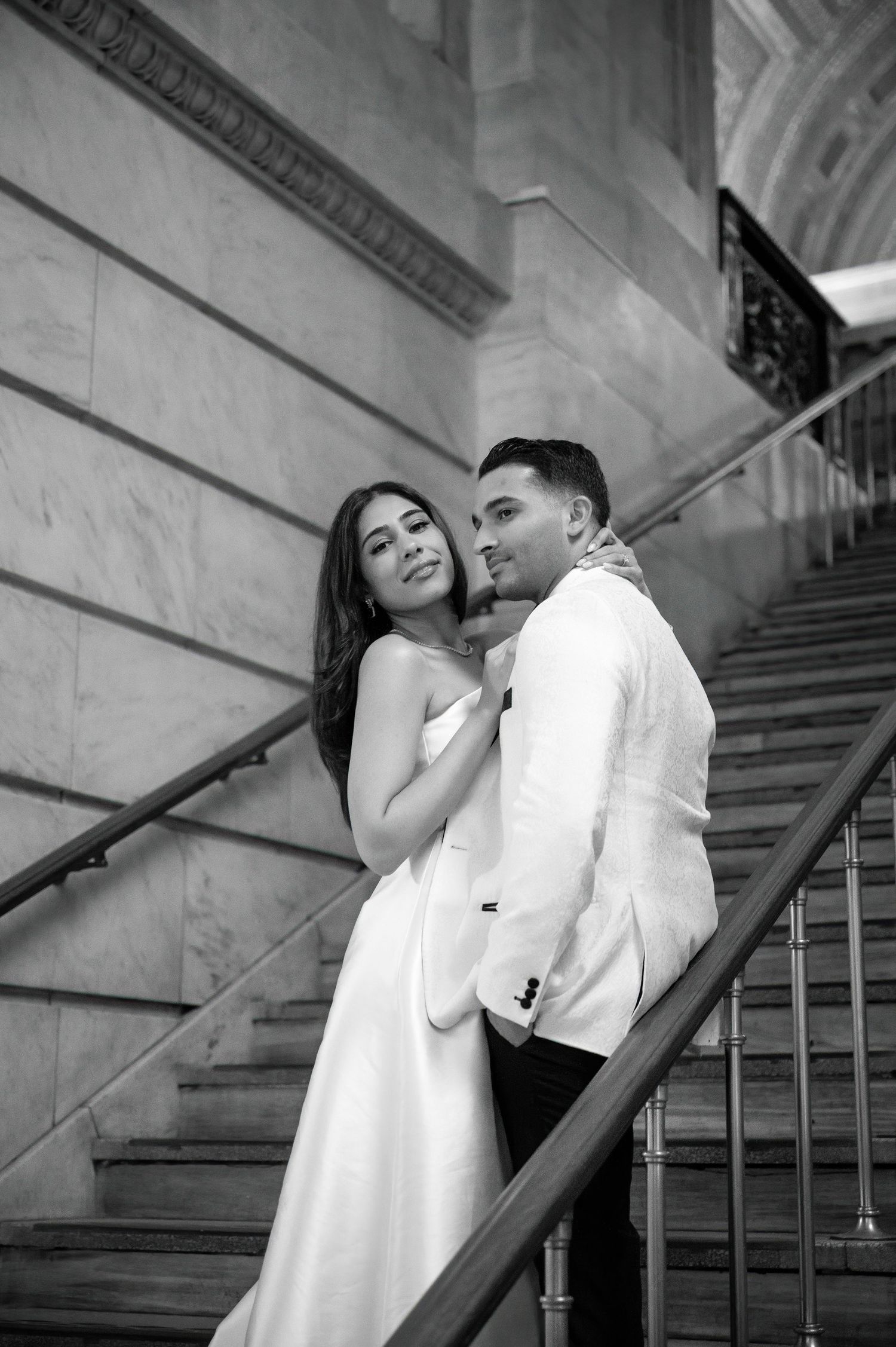A couple in formal attire poses romantically on an elegant marble staircase in a black and white photograph.