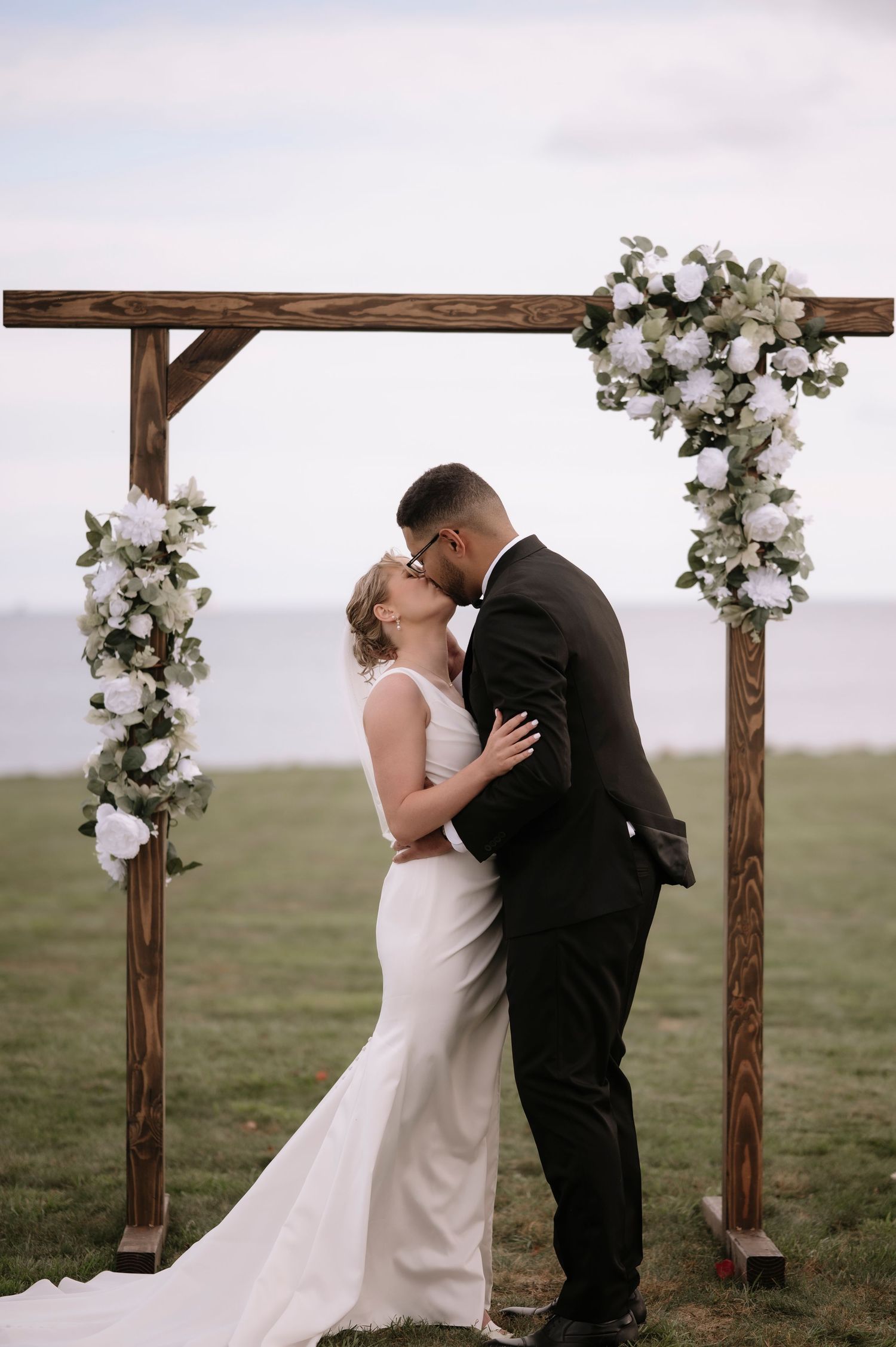 Newlyweds share a kiss under a rustic wooden arch adorned with white floral arrangements against an open sky.