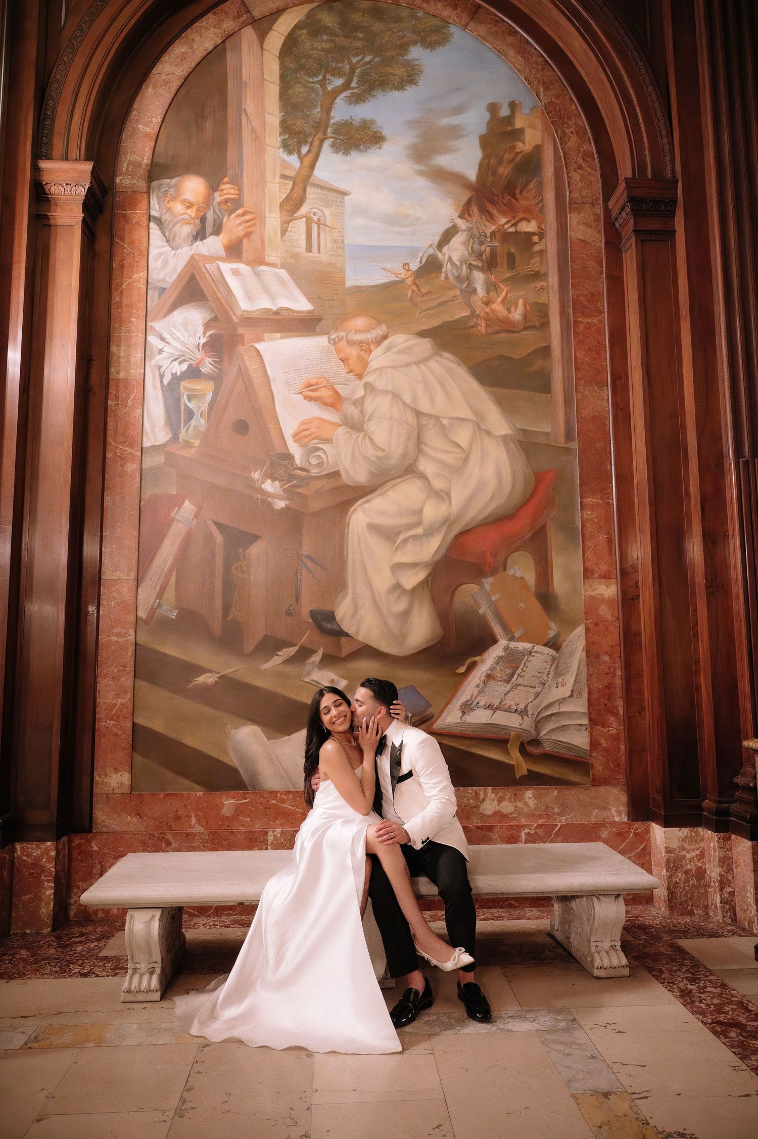 Wedding couple sits on stone bench beneath historic religious fresco in ornate archway setting.