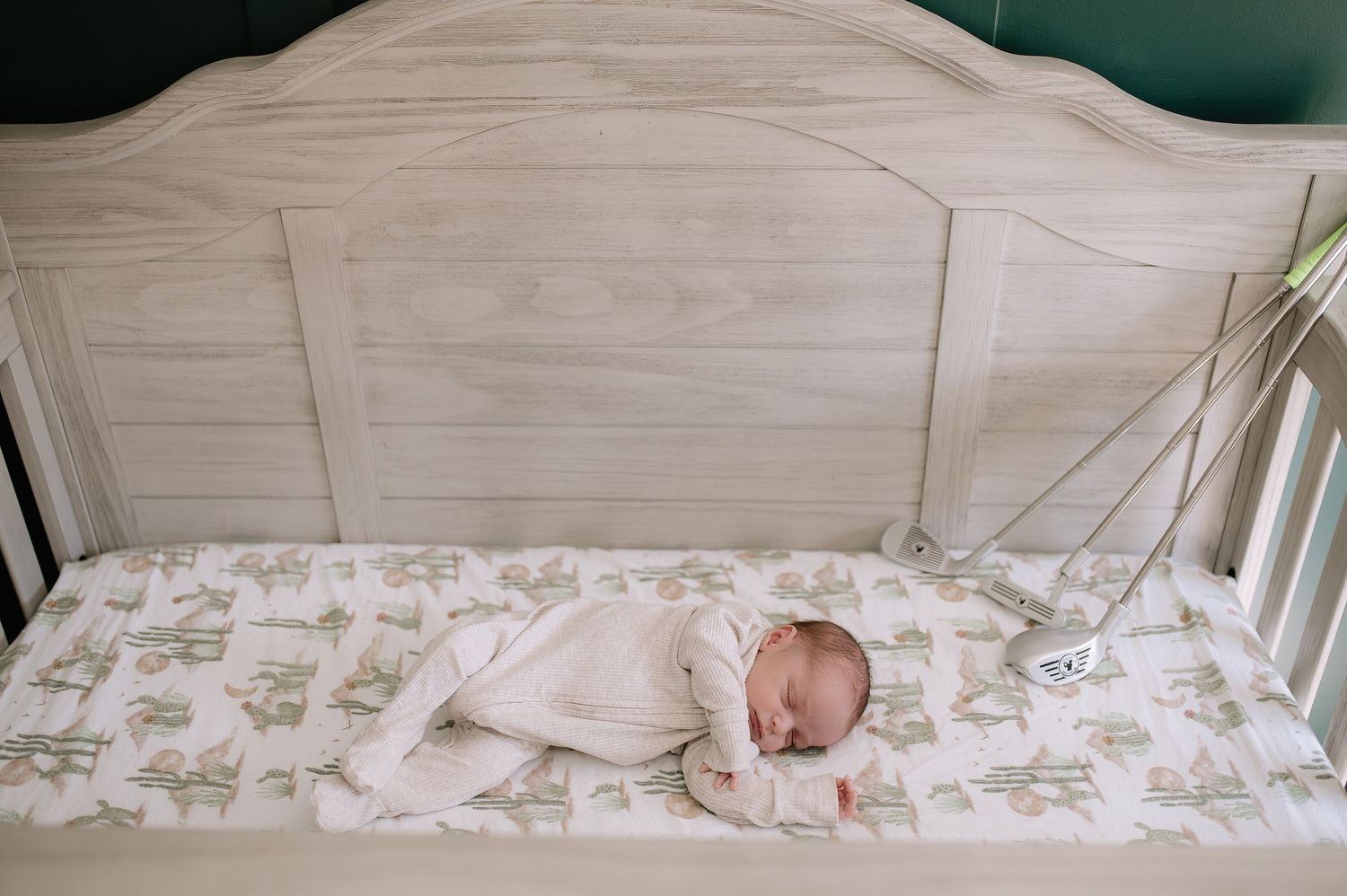 A sleeping newborn baby in white clothing lies on a patterned sheet in a wooden crib.