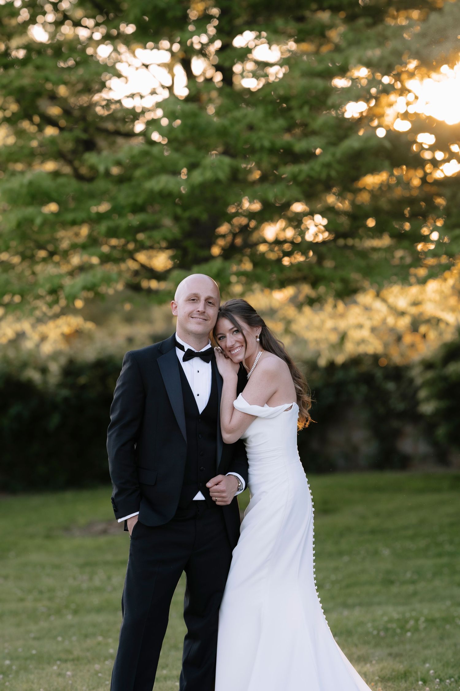 A bride in white and groom in tuxedo share a romantic outdoor moment during sunset at their wedding photoshoot.