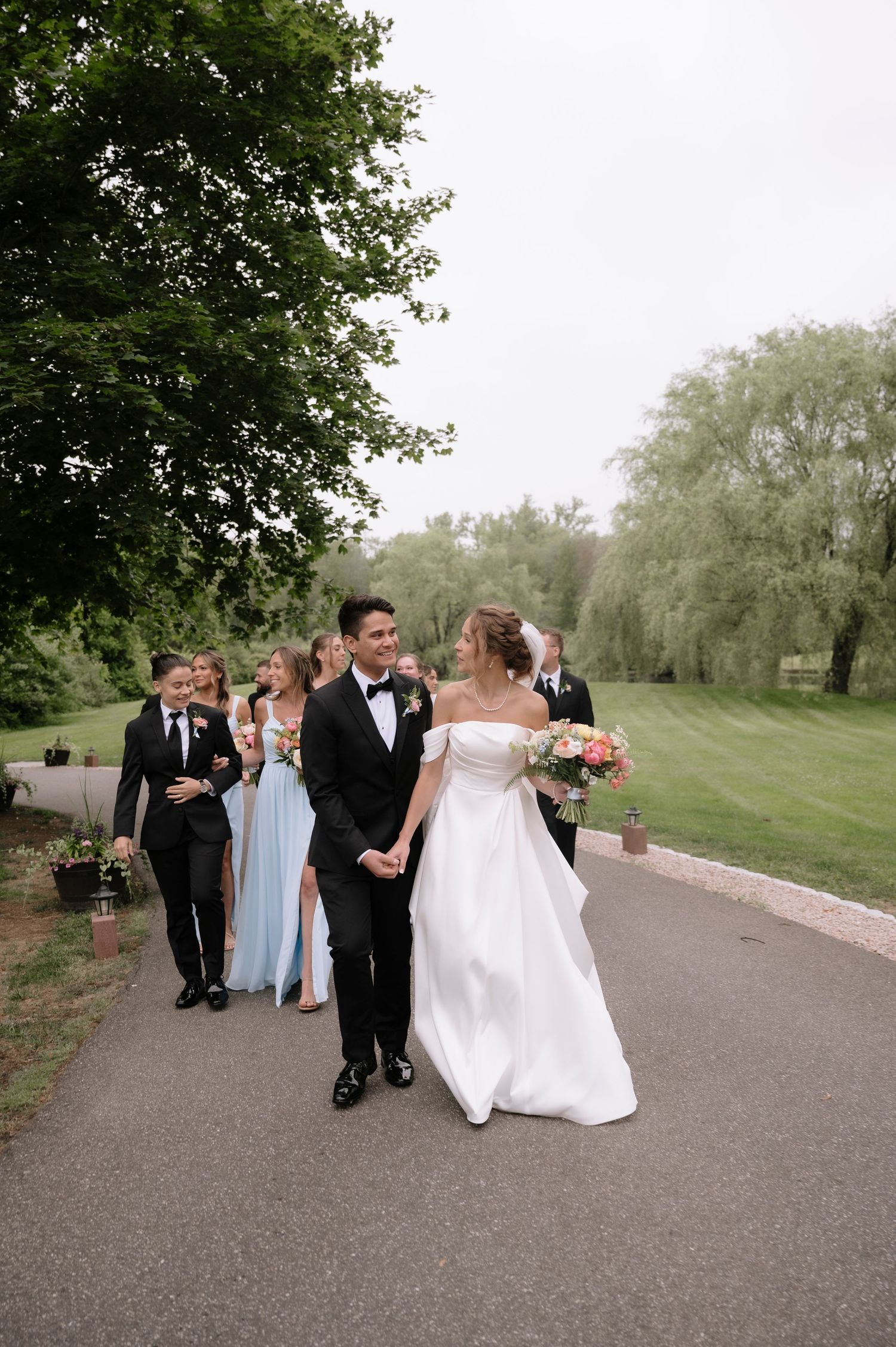 Wedding party walking and laughing together on a paved path surrounded by green trees and grass during an outdoor celebration.