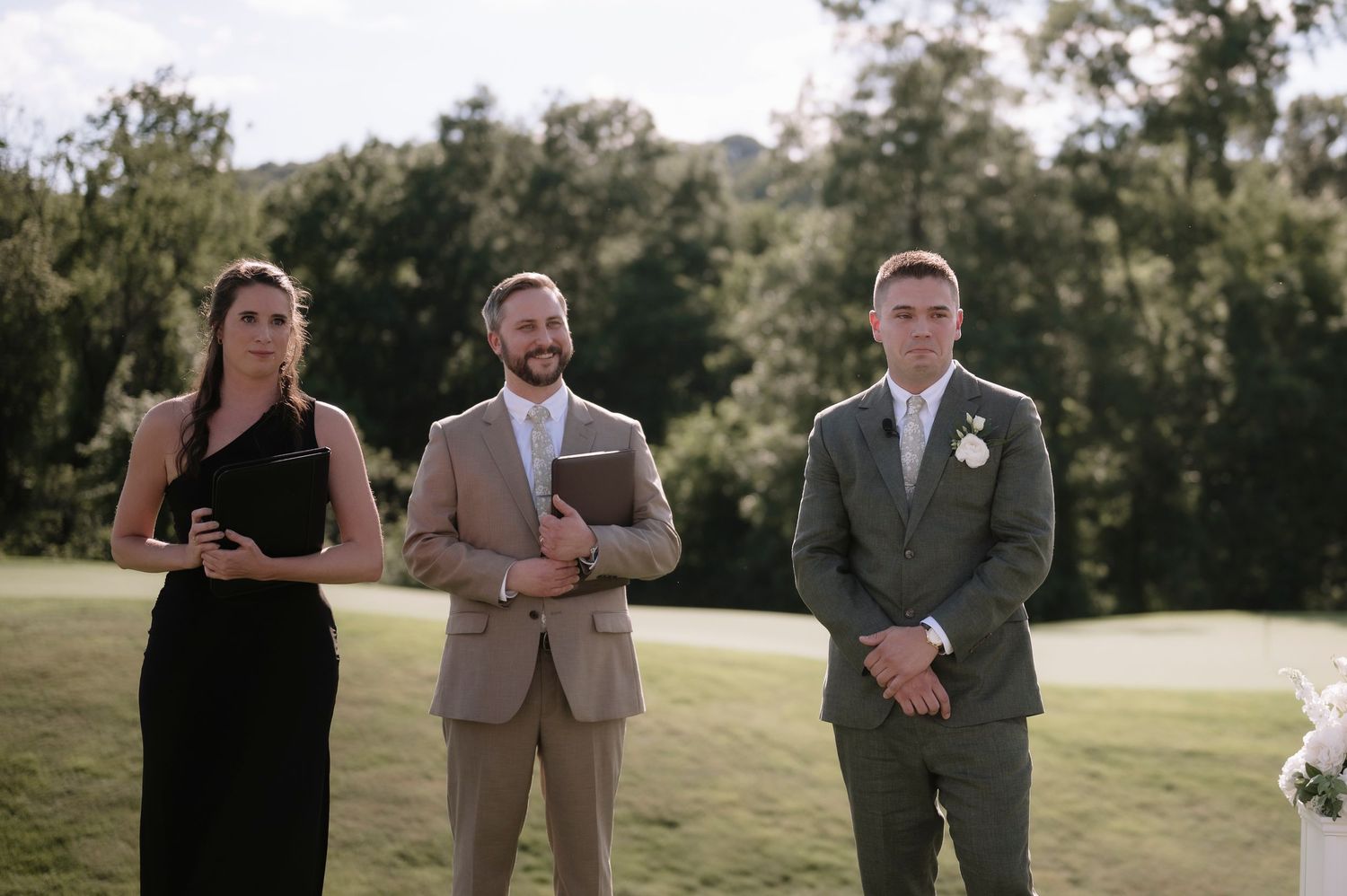Groom in green suit stands at outdoor wedding altar with officiant and witness on a lush golf course.