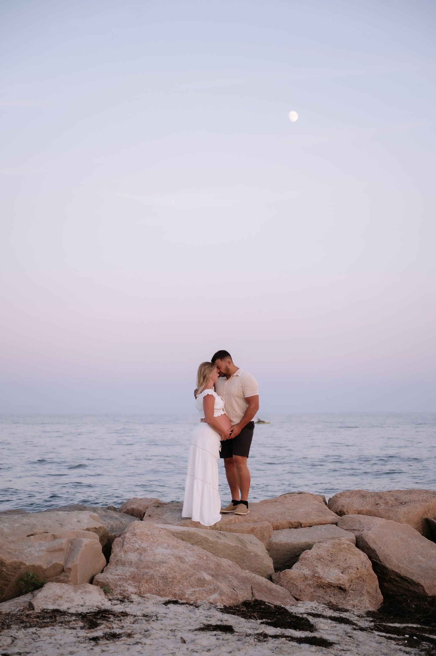 Couple sharing a romantic kiss on rocky shoreline at dusk with moon visible in pastel sky above calm ocean.