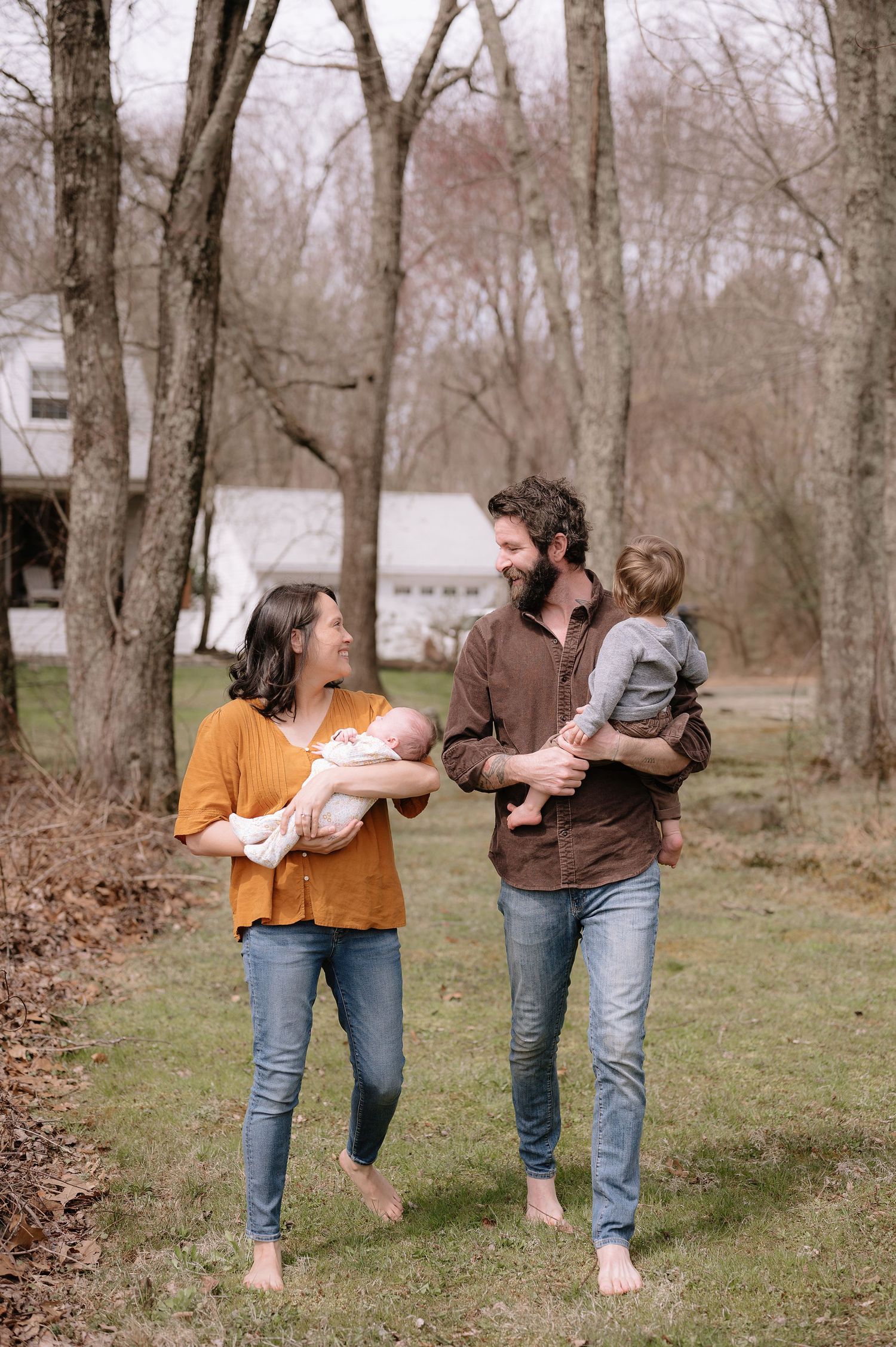 A barefoot couple walks outdoors holding their children against a backdrop of bare trees.