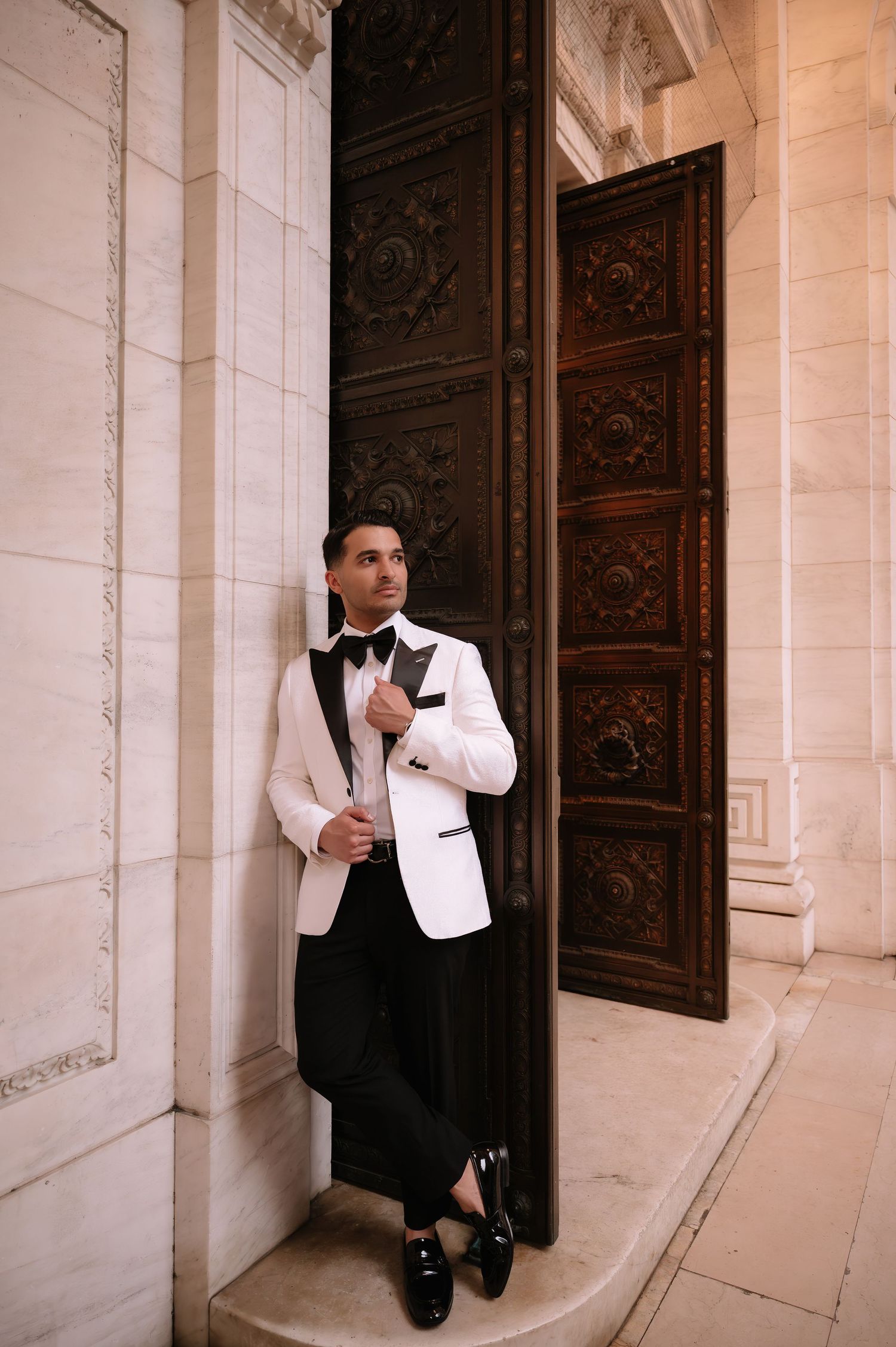 A person in a white tuxedo jacket and black bow tie leans against an ornate doorway with carved wooden details.