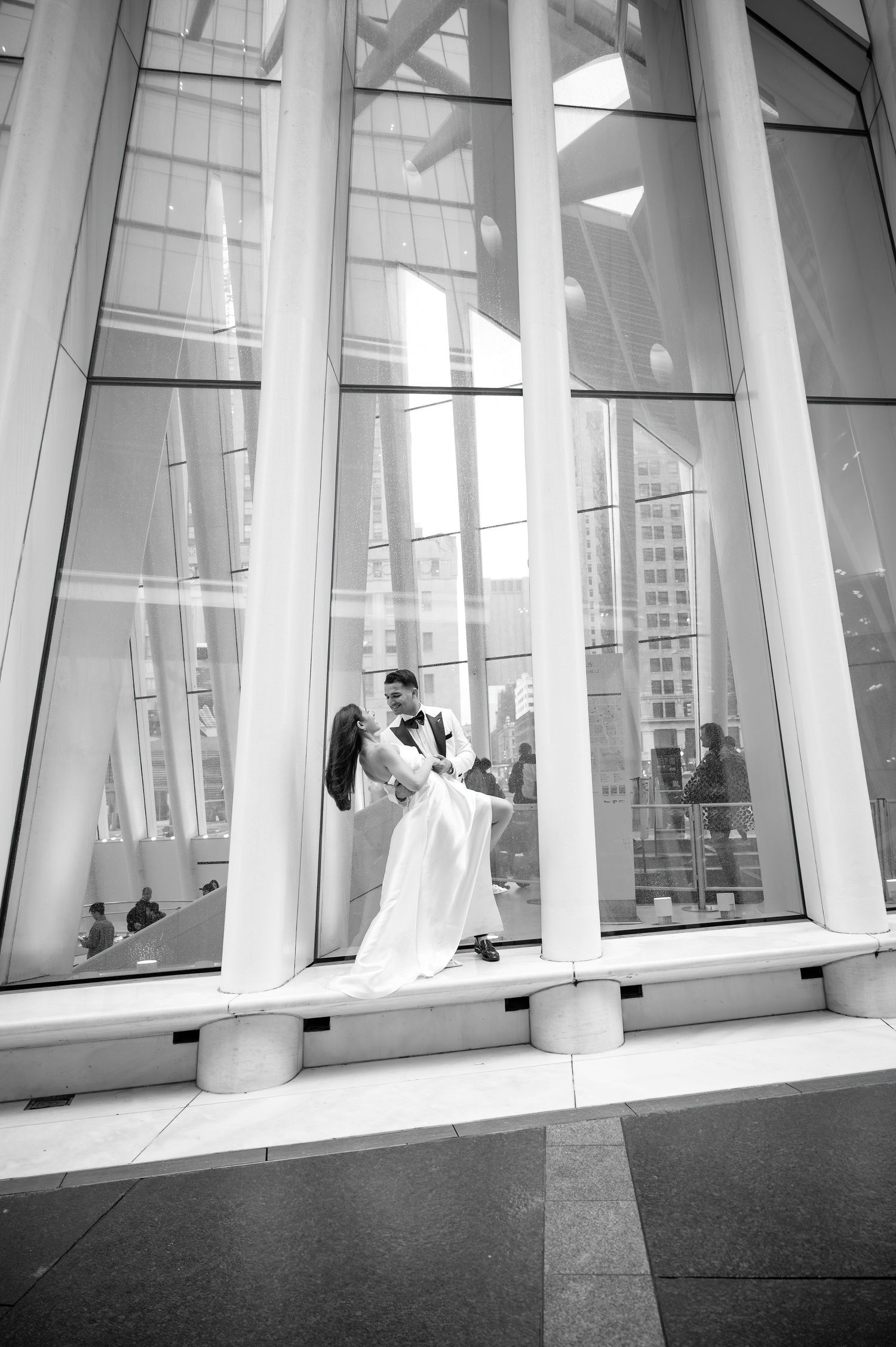 Black and white photo of couple in wedding attire posing between dramatic architectural columns.