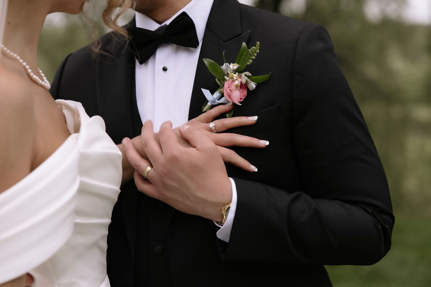 Close-up shot of hands touching a black tuxedo with pink floral boutonniere at formal wedding event.