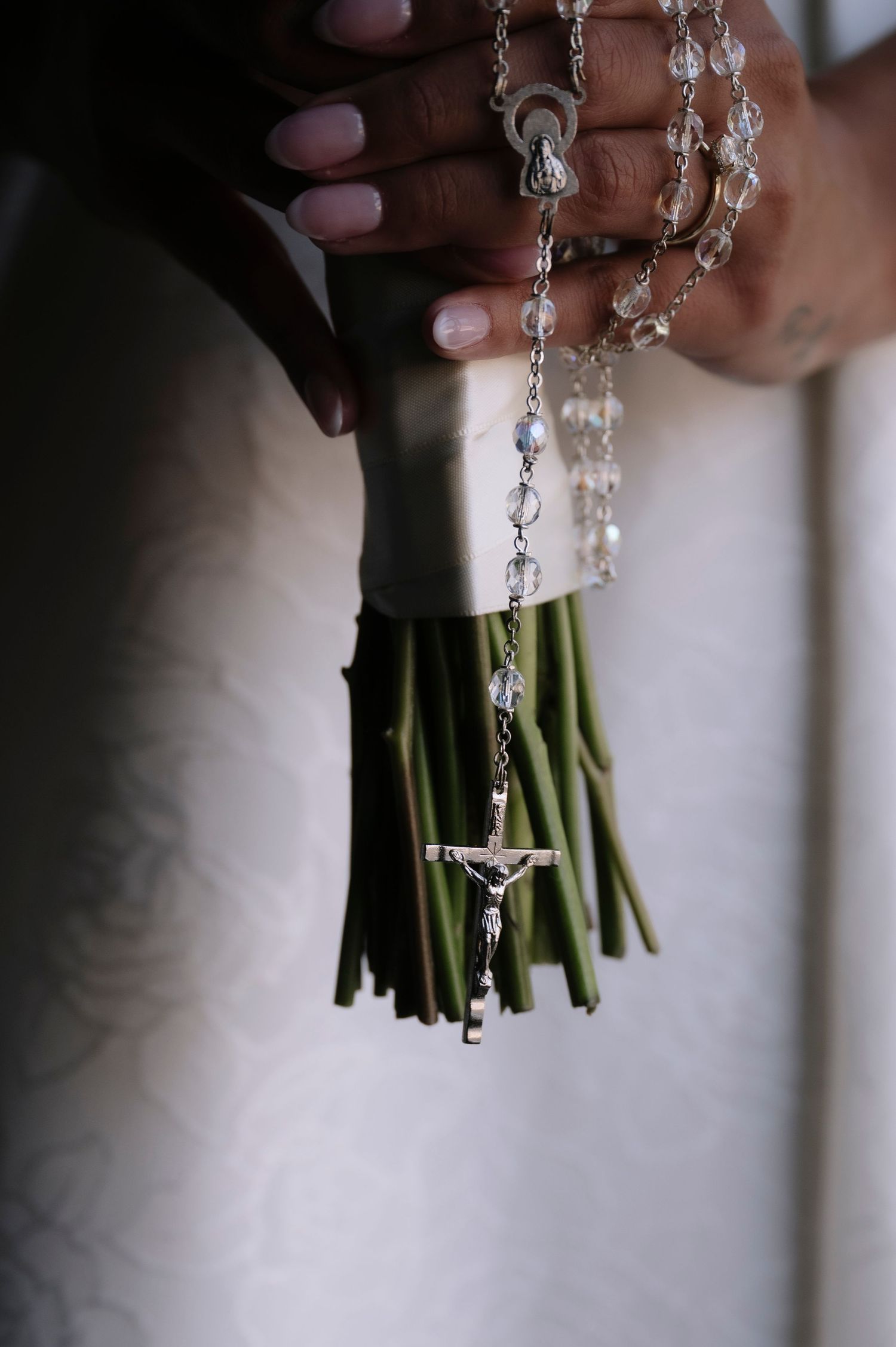 Hands holding a bridal bouquet with a silver rosary and crucifix draped over green stems.