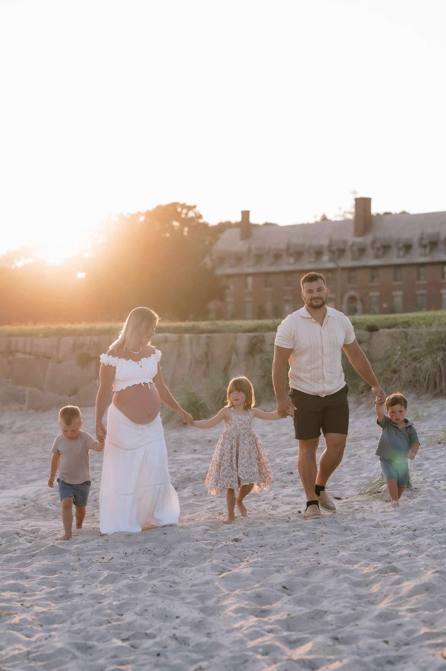 Family of five walking on sandy beach at golden sunset, pregnant mother in white dress holding hands with children.