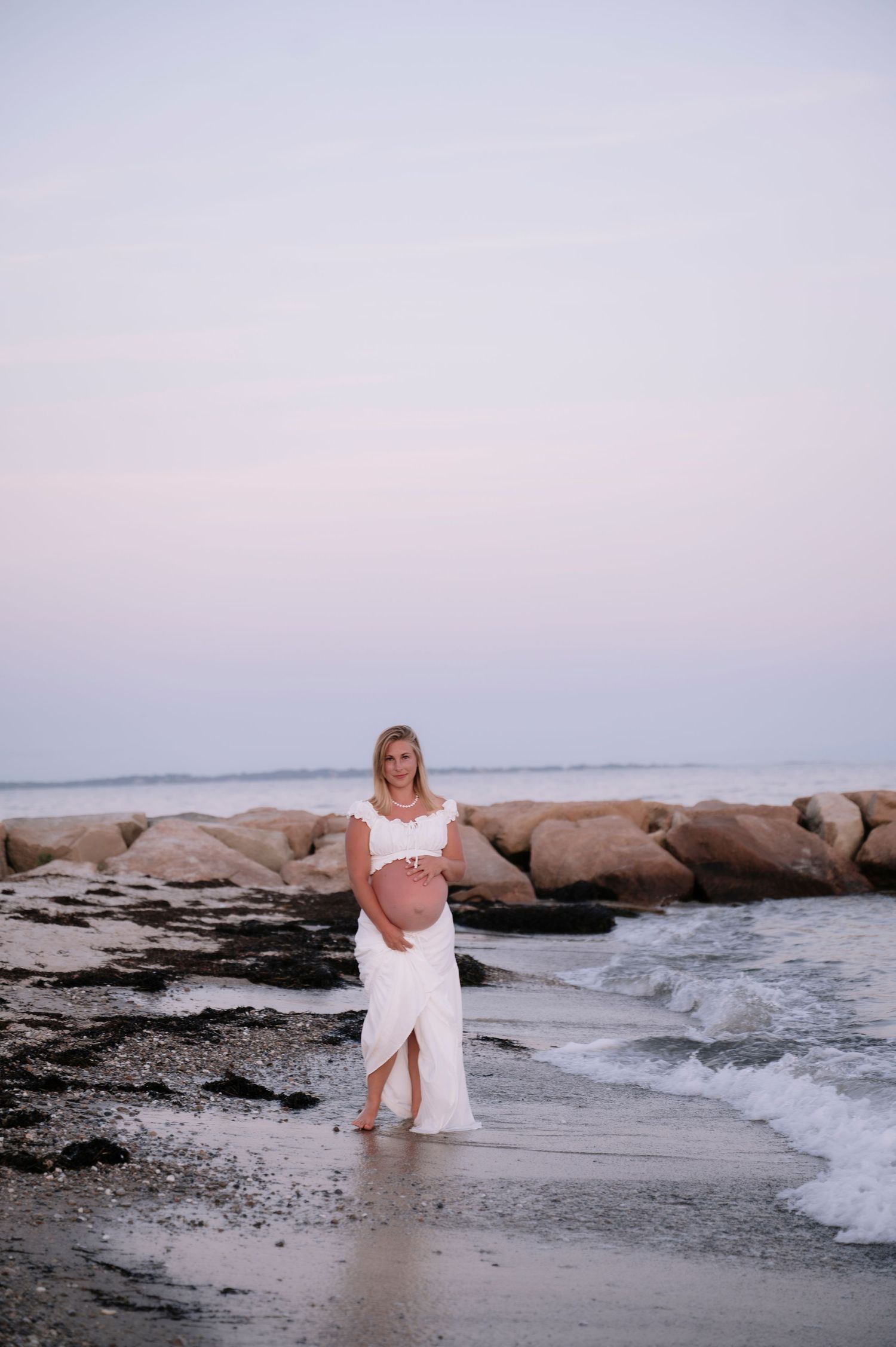 Pregnant woman in white dress cradling baby bump on rocky beach at sunset, waves gently washing ashore.