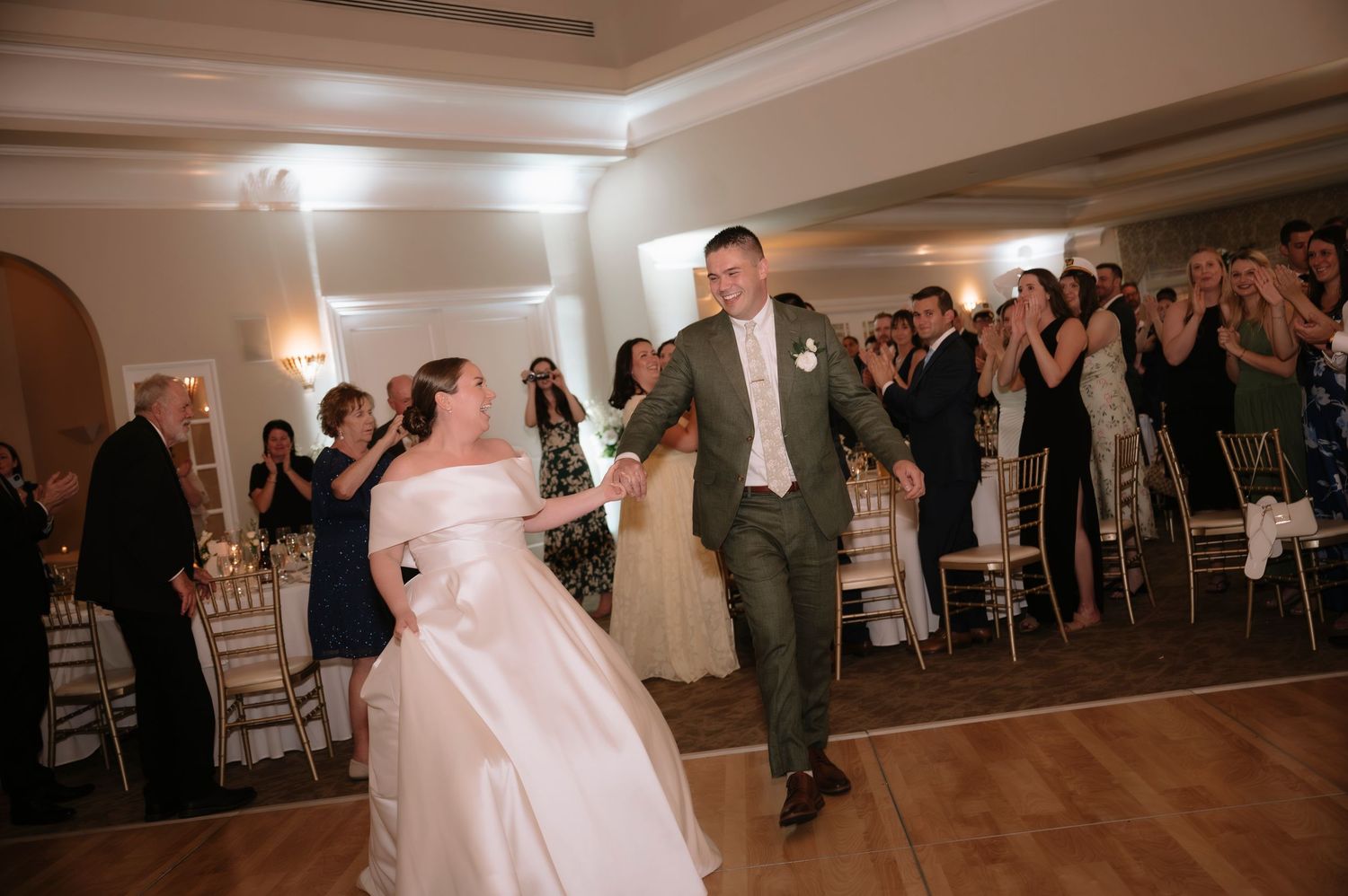 Bride in white off-shoulder gown and groom in green suit share first dance at elegant wedding reception.