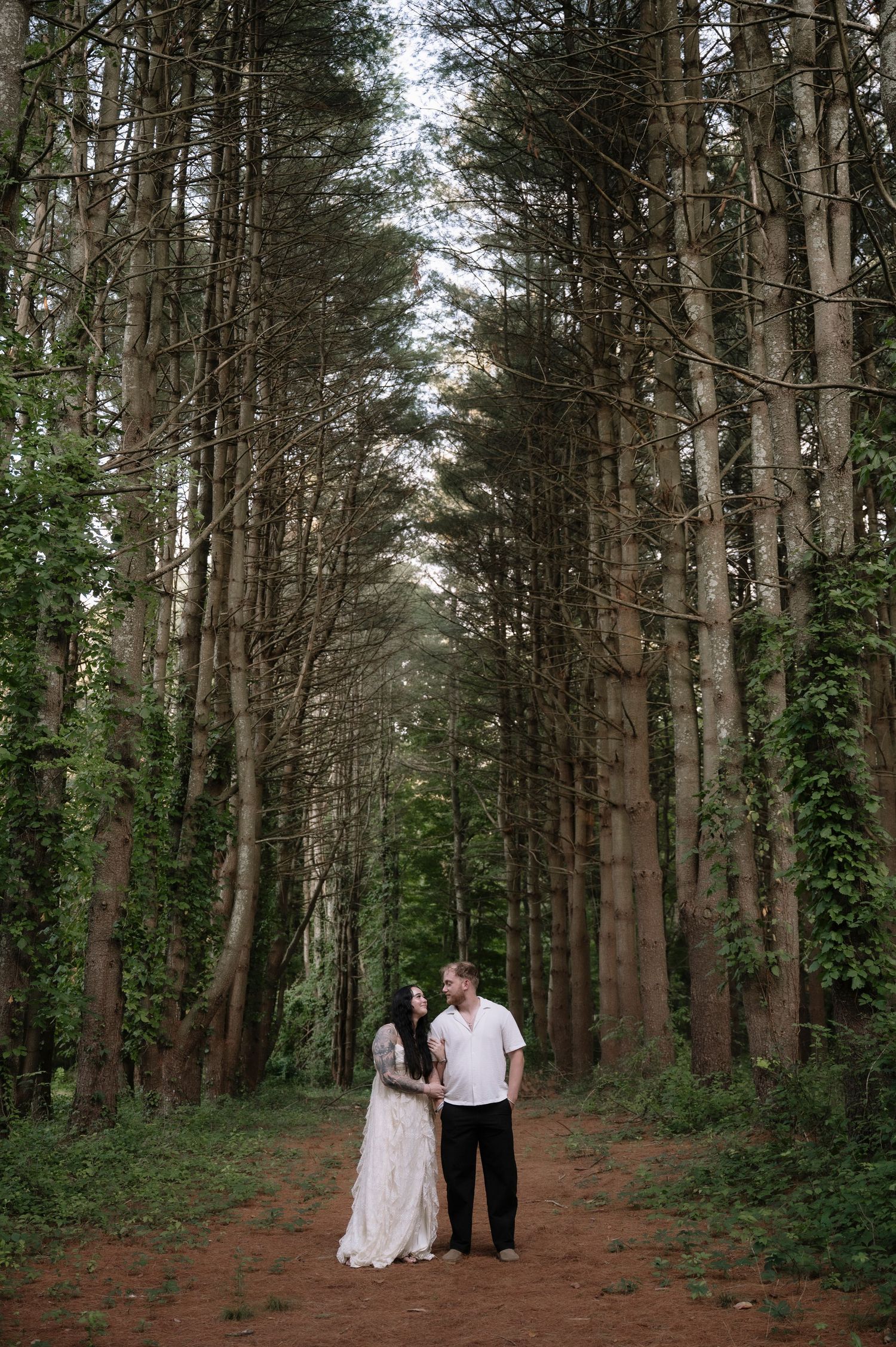 A couple stands hand-in-hand on a forest path surrounded by tall trees, the woman in a white gown and man in casual attire.