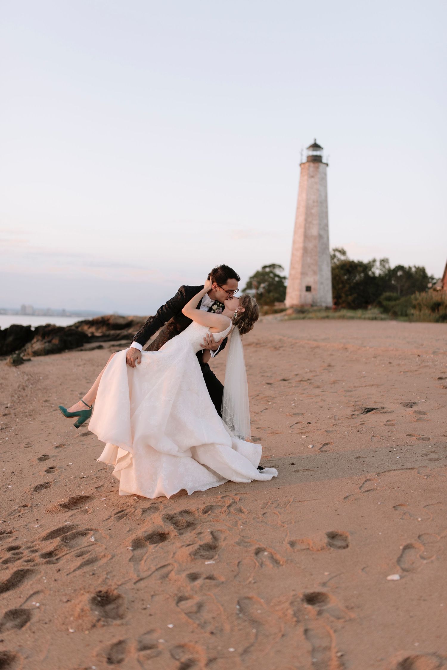 Romantic sunset photo of newlyweds embracing near a lighthouse on a sandy beach.