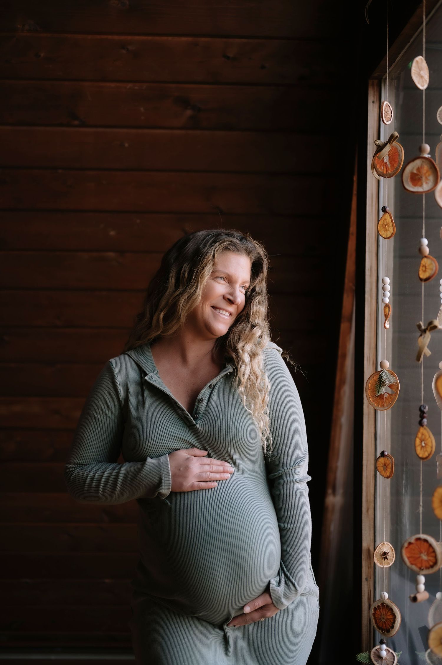 Expecting mother in sage green dress stands by window with dried citrus garland decorations.