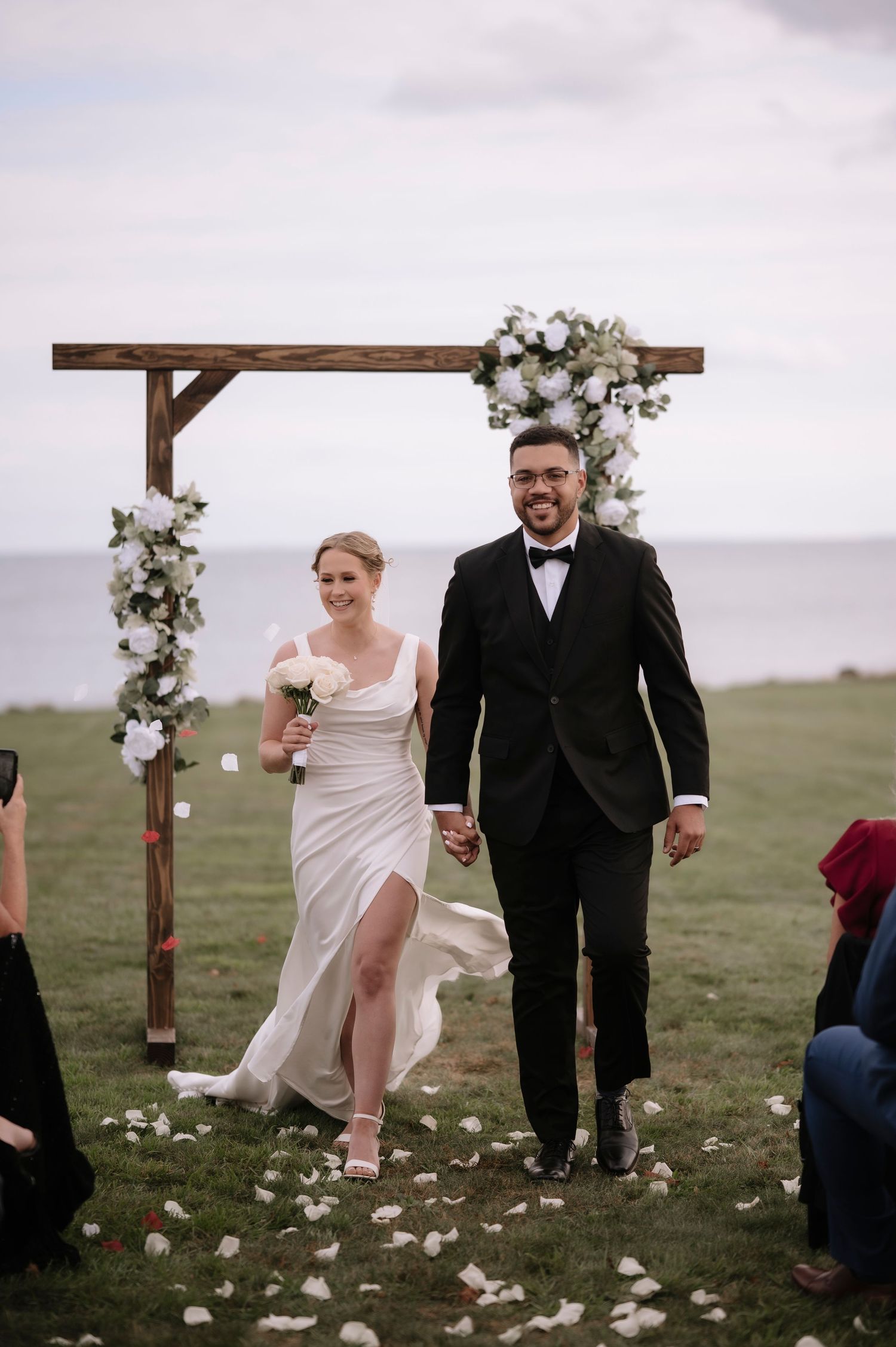 A wedding couple walks through scattered flower petals under a rustic wooden arch decorated with white flowers.