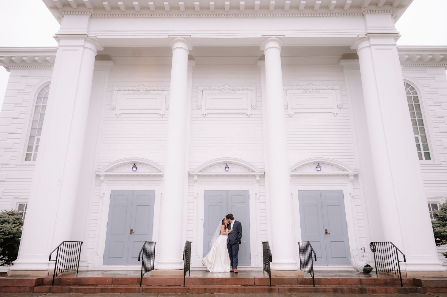 Wide angle wedding portrait on church steps with white columns and gray doors.