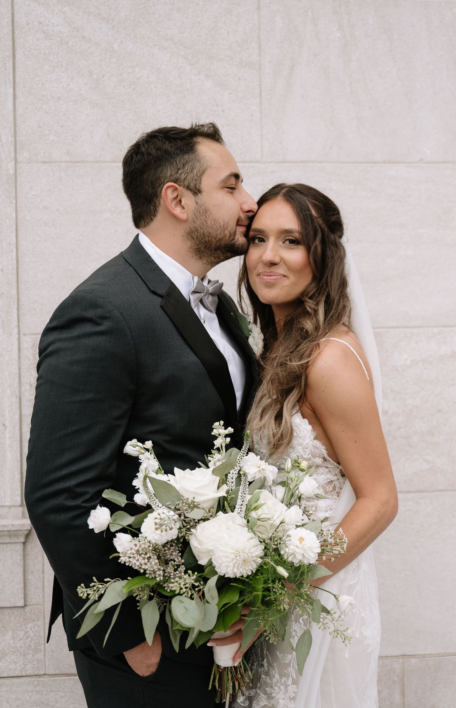Groom kisses bride while holding white floral bouquet against stone wall.