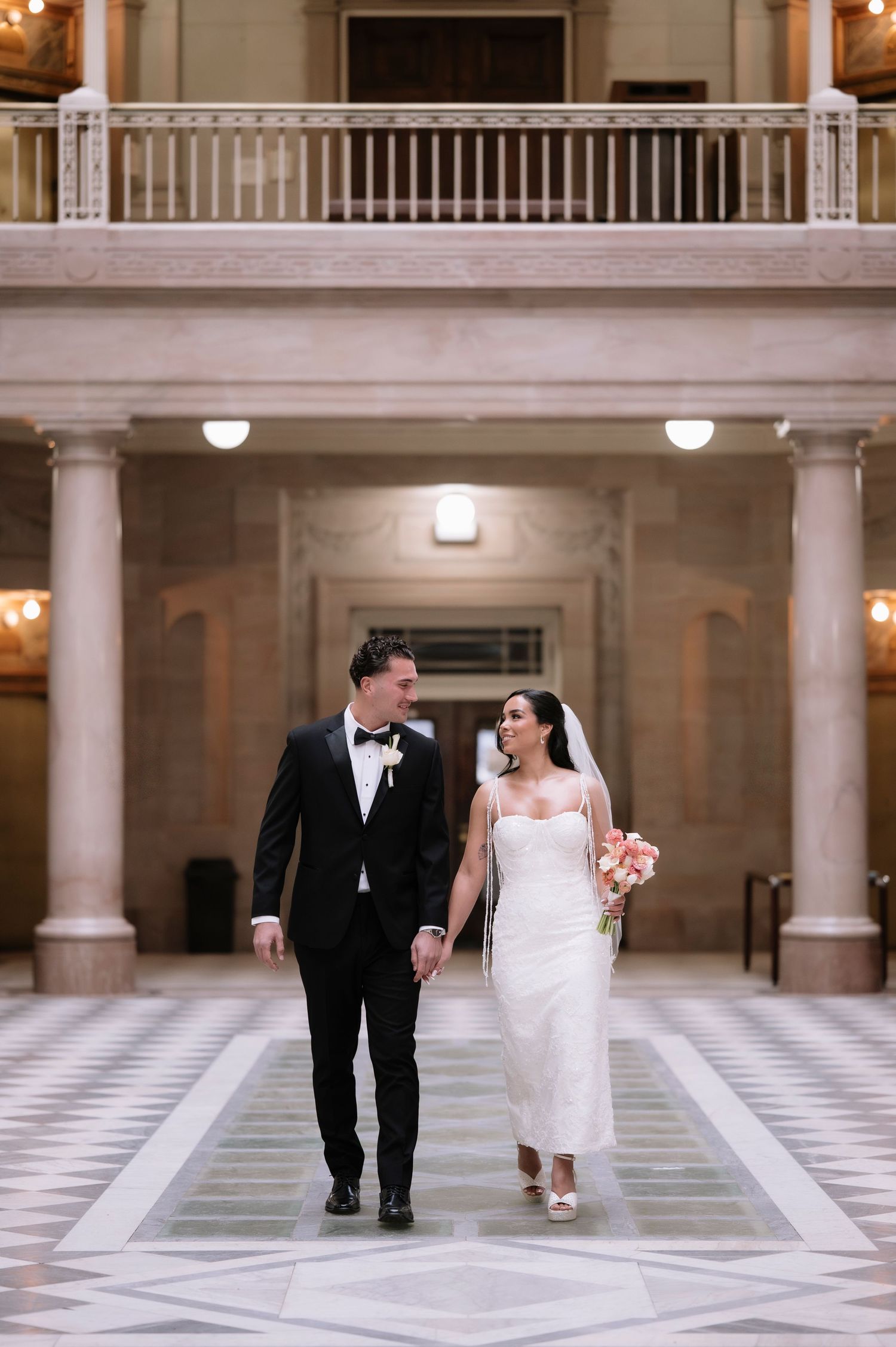 Bride and groom walk hand-in-hand through an elegant marble hall, she in a white gown holding pink blooms, he in a black tuxedo.