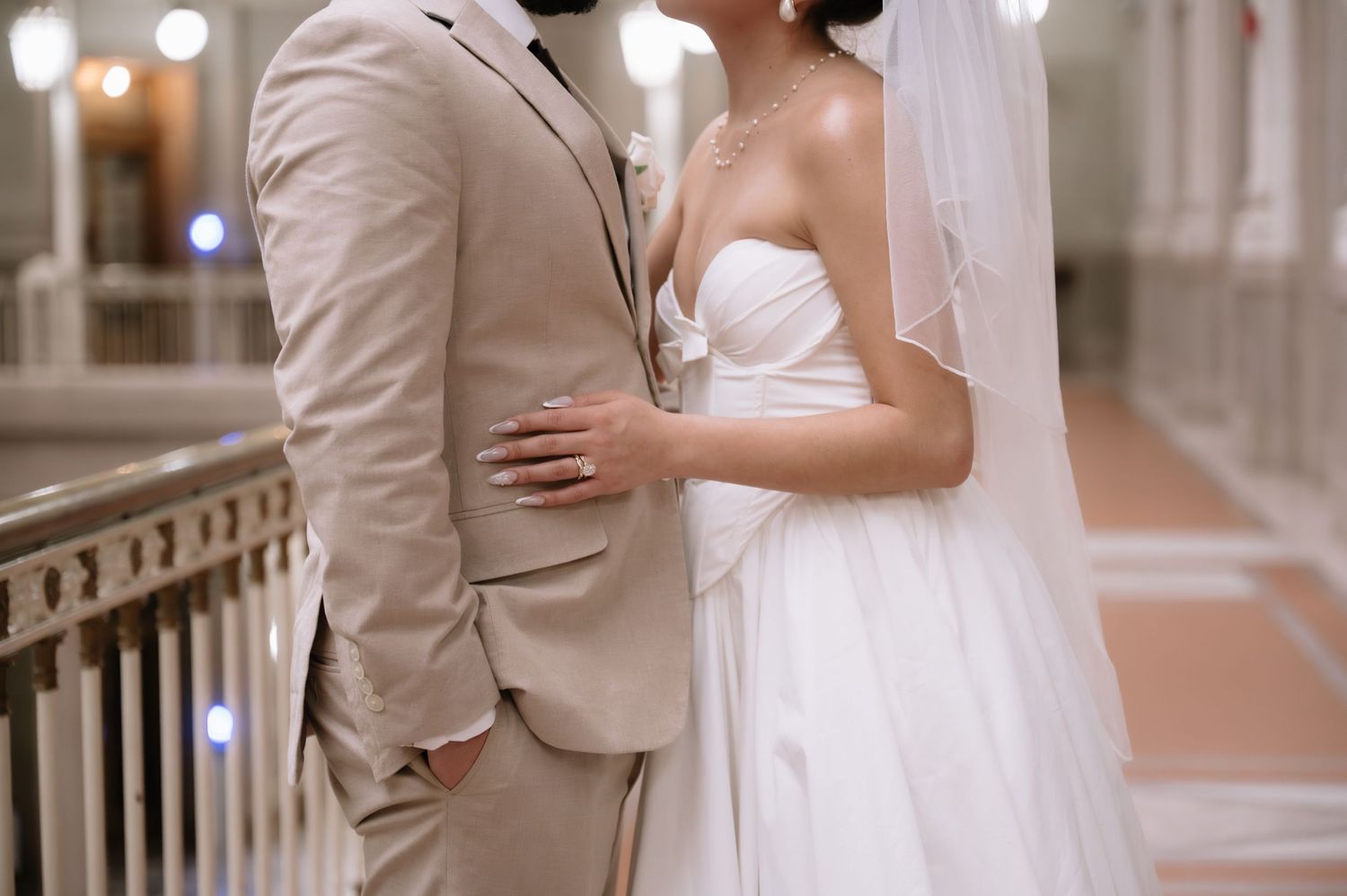 Elegant couple in beige and white formal attire pose together in a bright indoor venue with decorative railings.