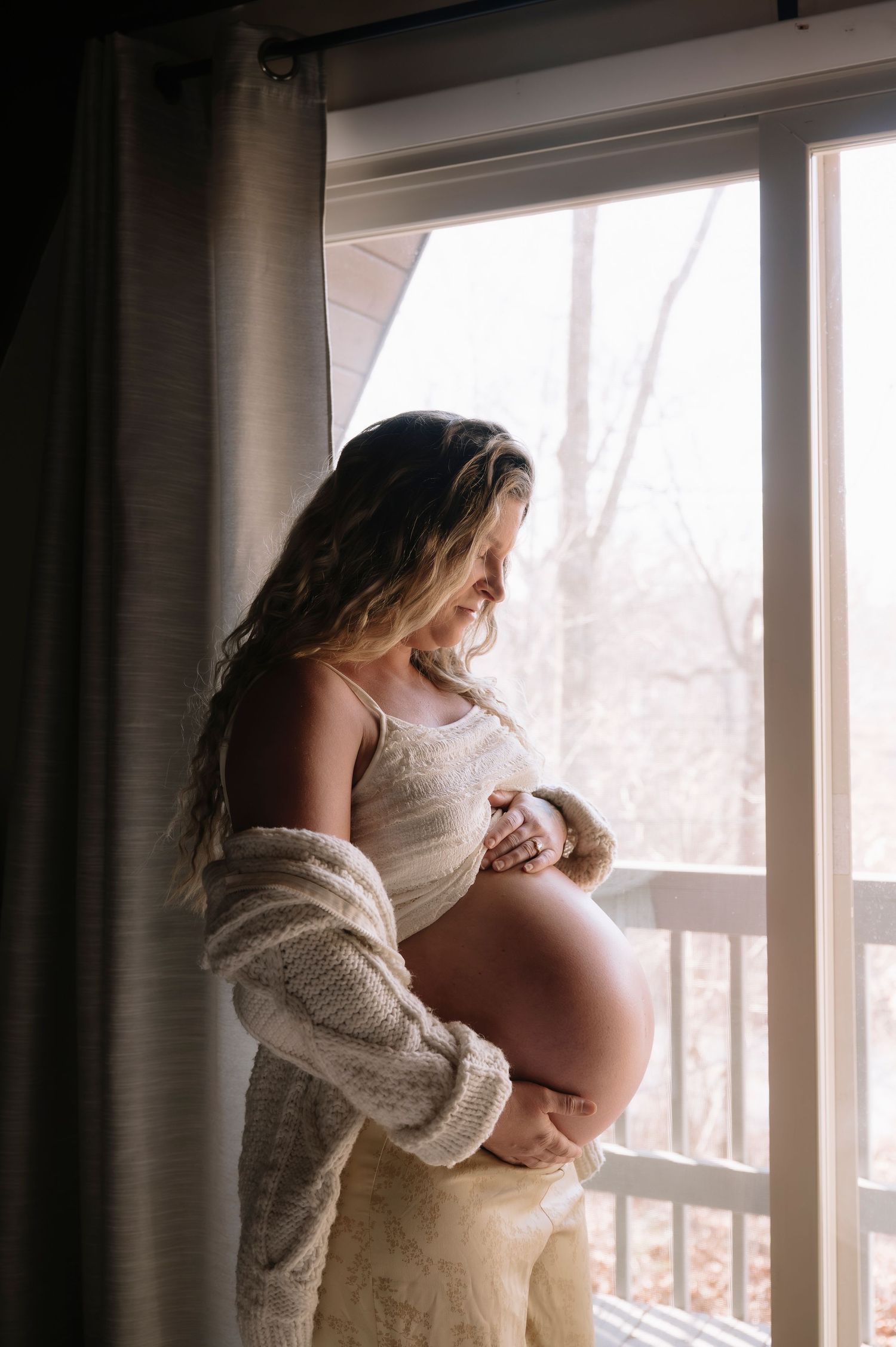 Artistic maternity portraits by a bright window showing a person in a cream sweater.