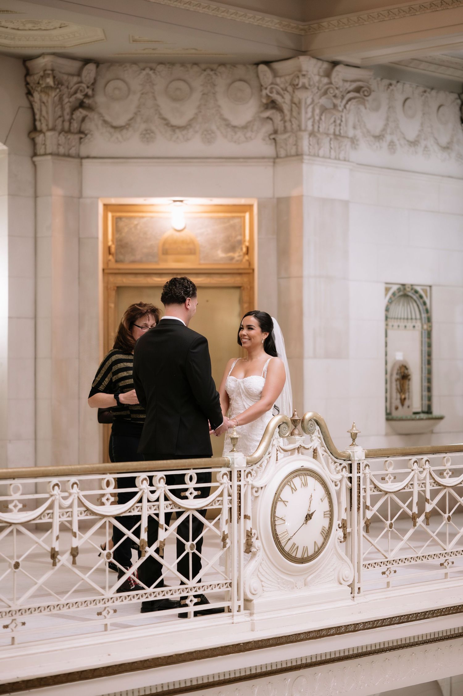 Couple exchanges vows beside elegant clock railing in grand hall.