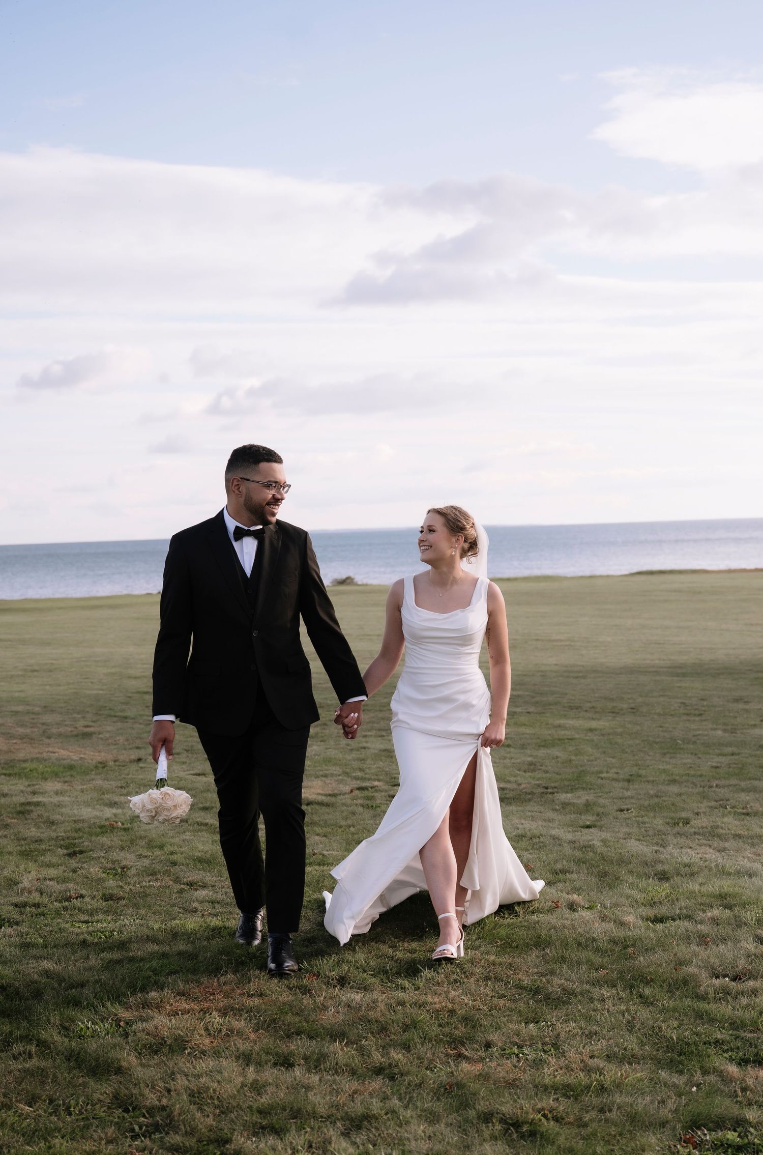 Couple walks hand in hand across grassy seaside cliff during sunset wedding photos.
