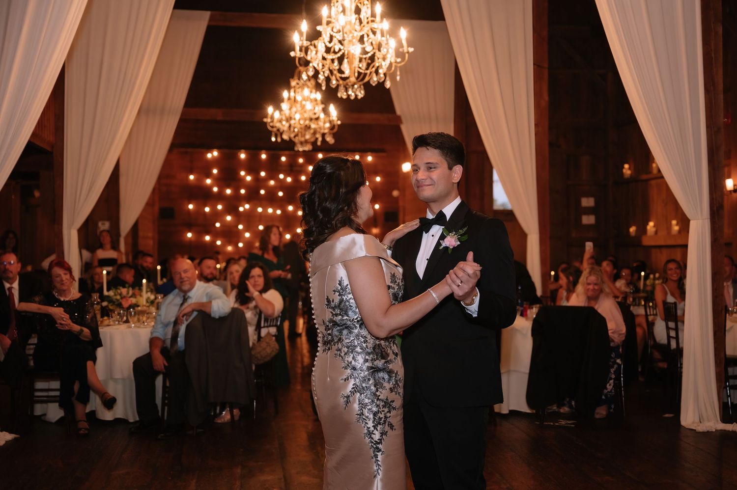 Bride and groom share first dance at elegant wedding reception with chandeliers and draped white curtains.