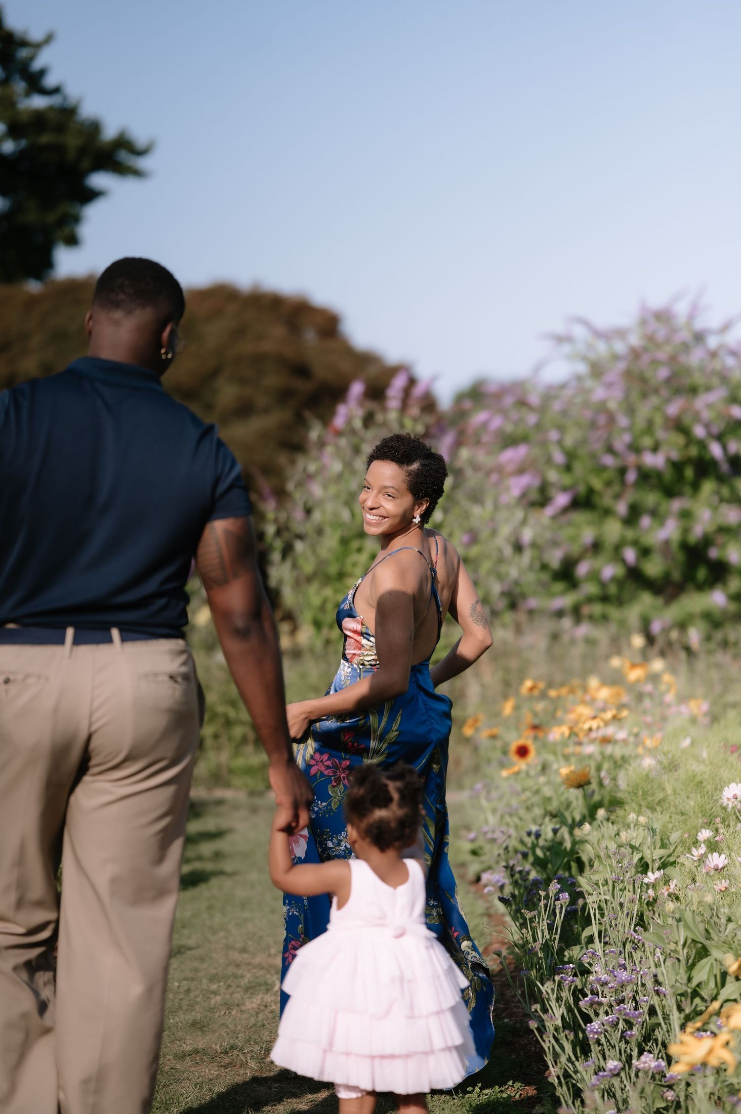 A joyful family of three walks through a colorful garden, with a smiling mother in a floral dress looking back at the camera.