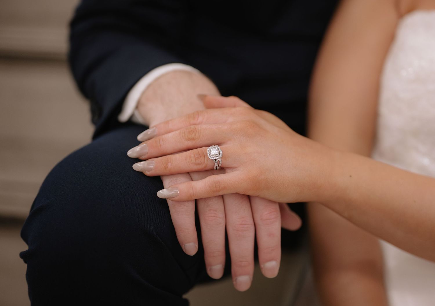 Close-up of hands with engagement ring resting on dark suit fabric.