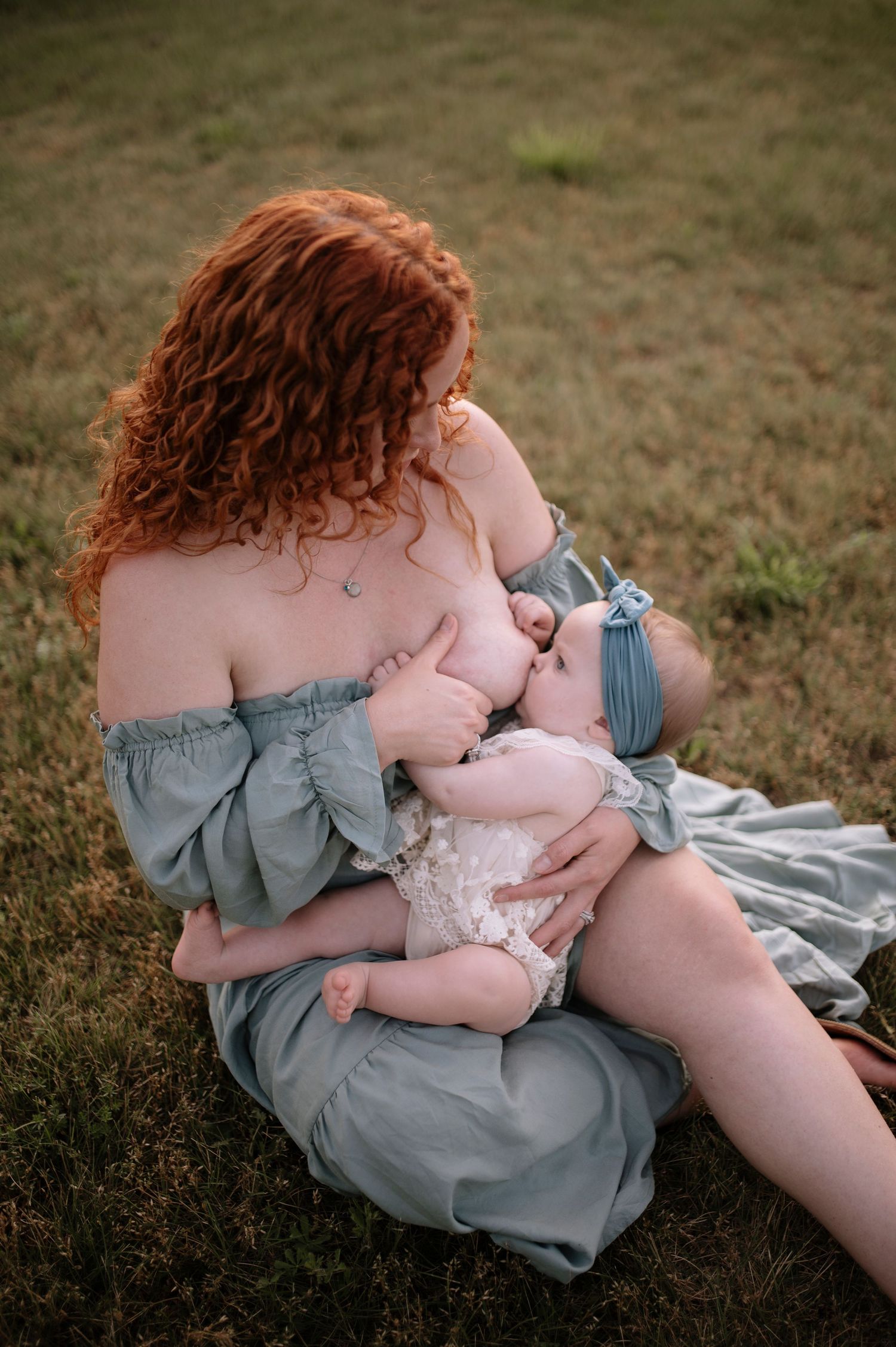A mother in a soft blue dress breastfeeds her baby while sitting on a blanket in a grassy field during golden hour.