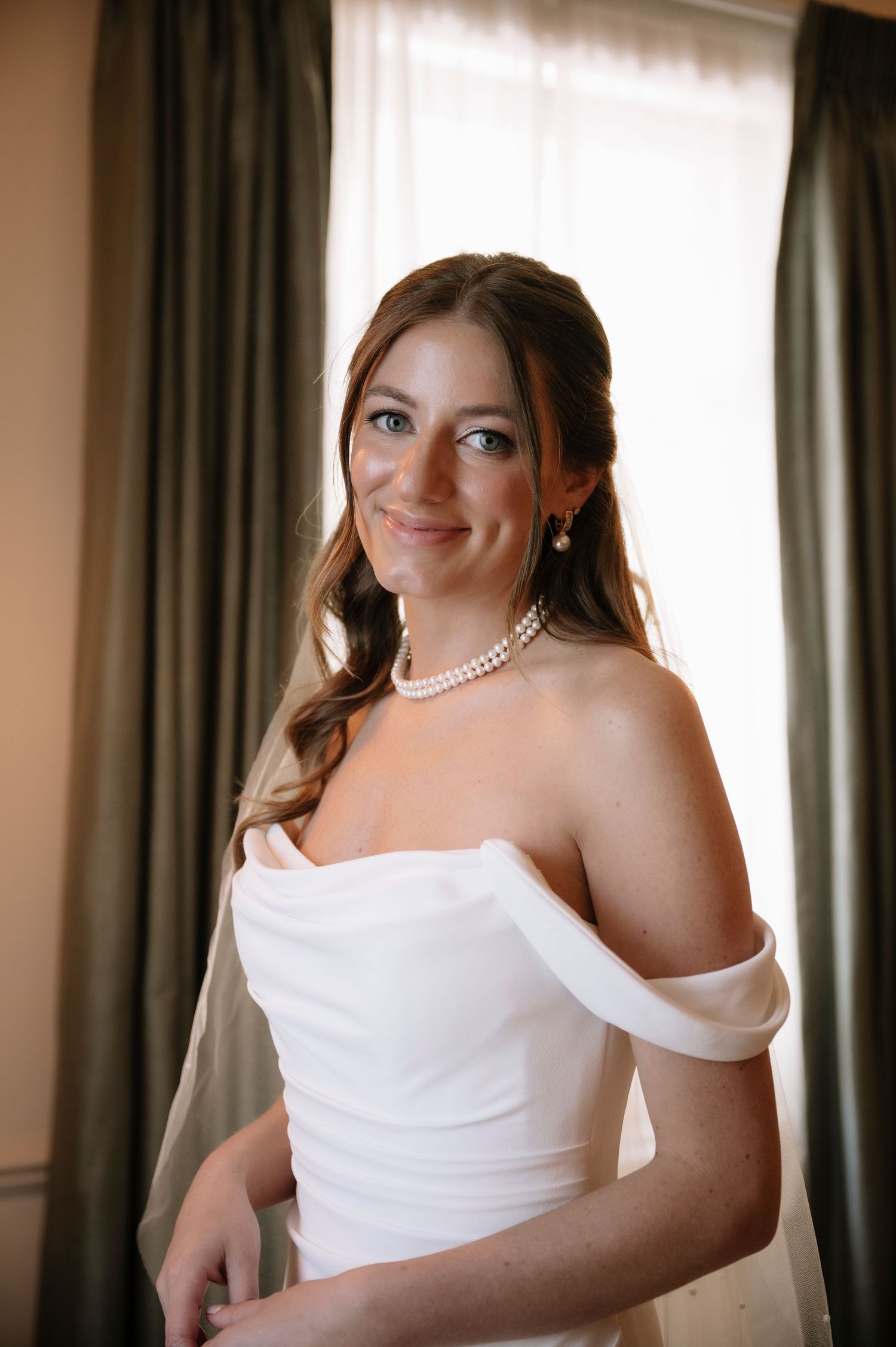 Elegant bride in white off-shoulder gown wearing pearl necklace poses by window with natural lighting.