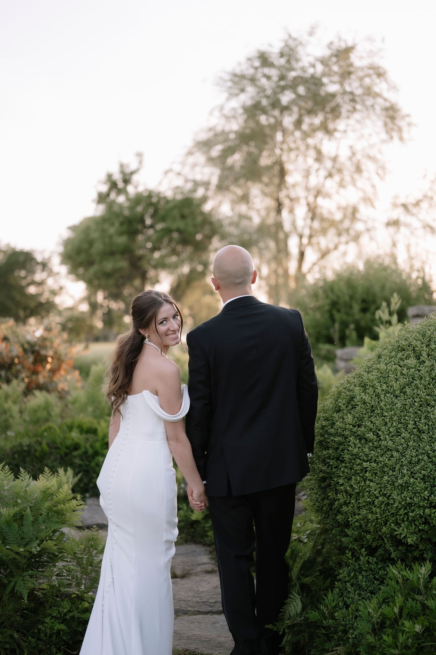 A couple in wedding attire walks along a garden path during golden hour surrounded by lush greenery.