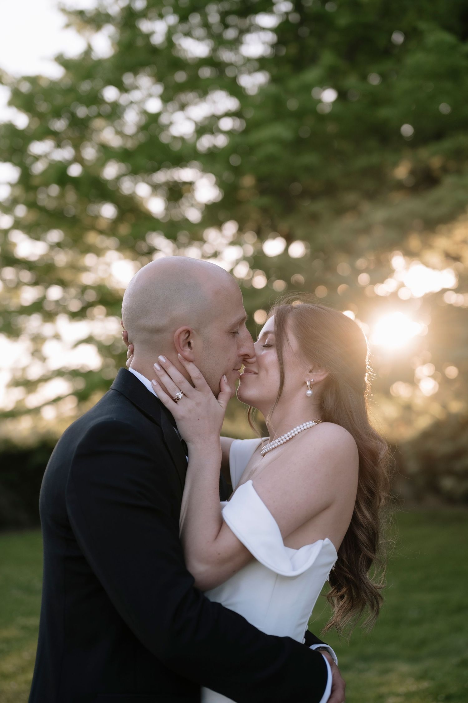 A couple shares a romantic kiss at sunset with sunlight streaming through trees in the background during their wedding.