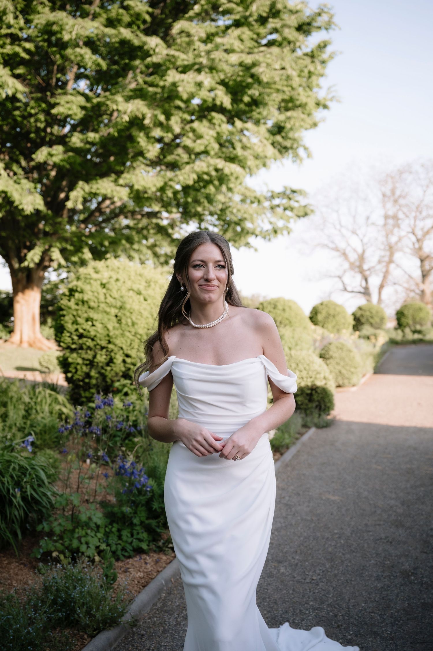 A person in a flowing white wedding dress stands serenely in a garden pathway surrounded by lush greenery and flowers.