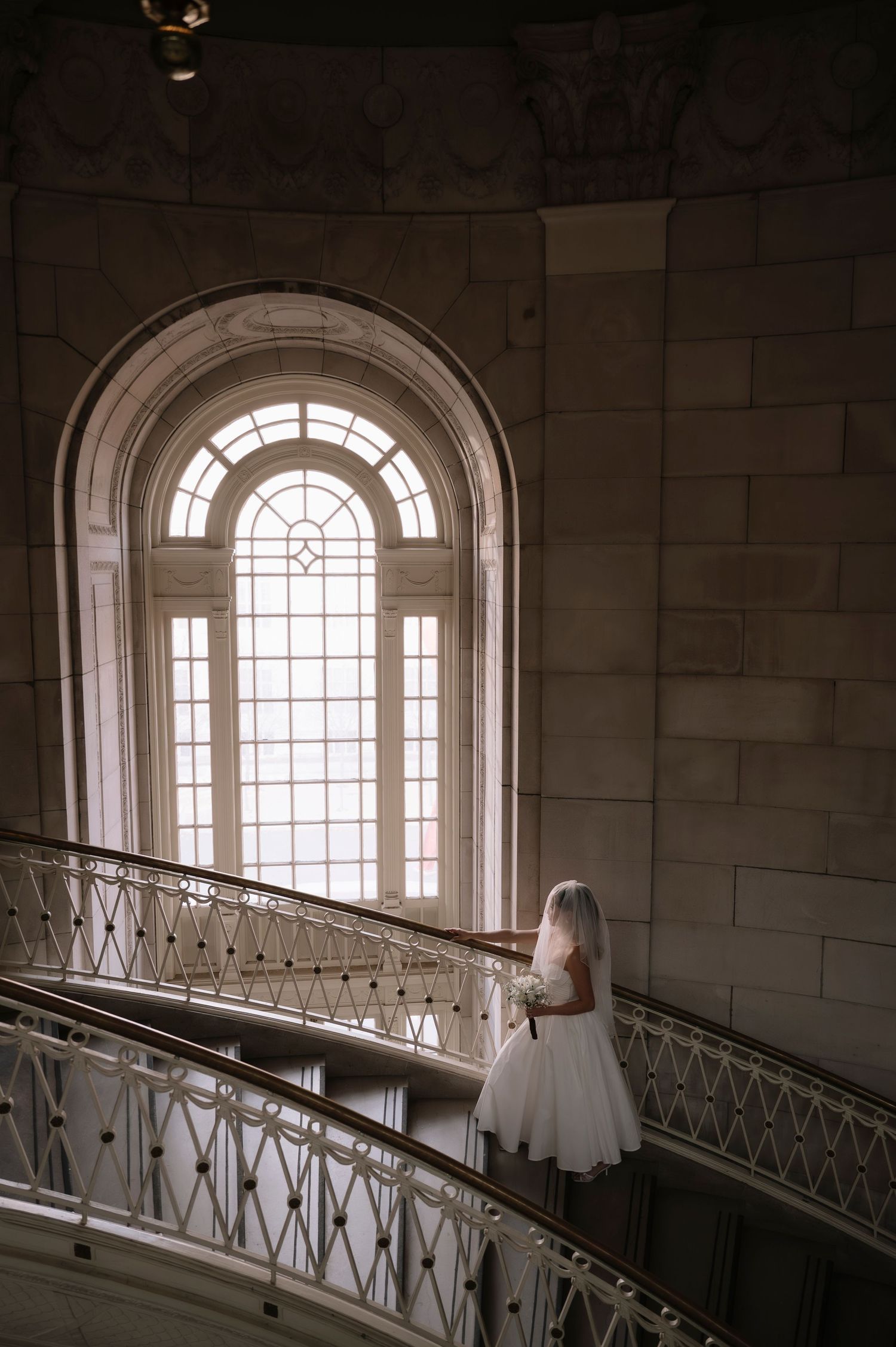 Architectural detail of a curved stone staircase with arched windows and natural light streaming through in a historic building.