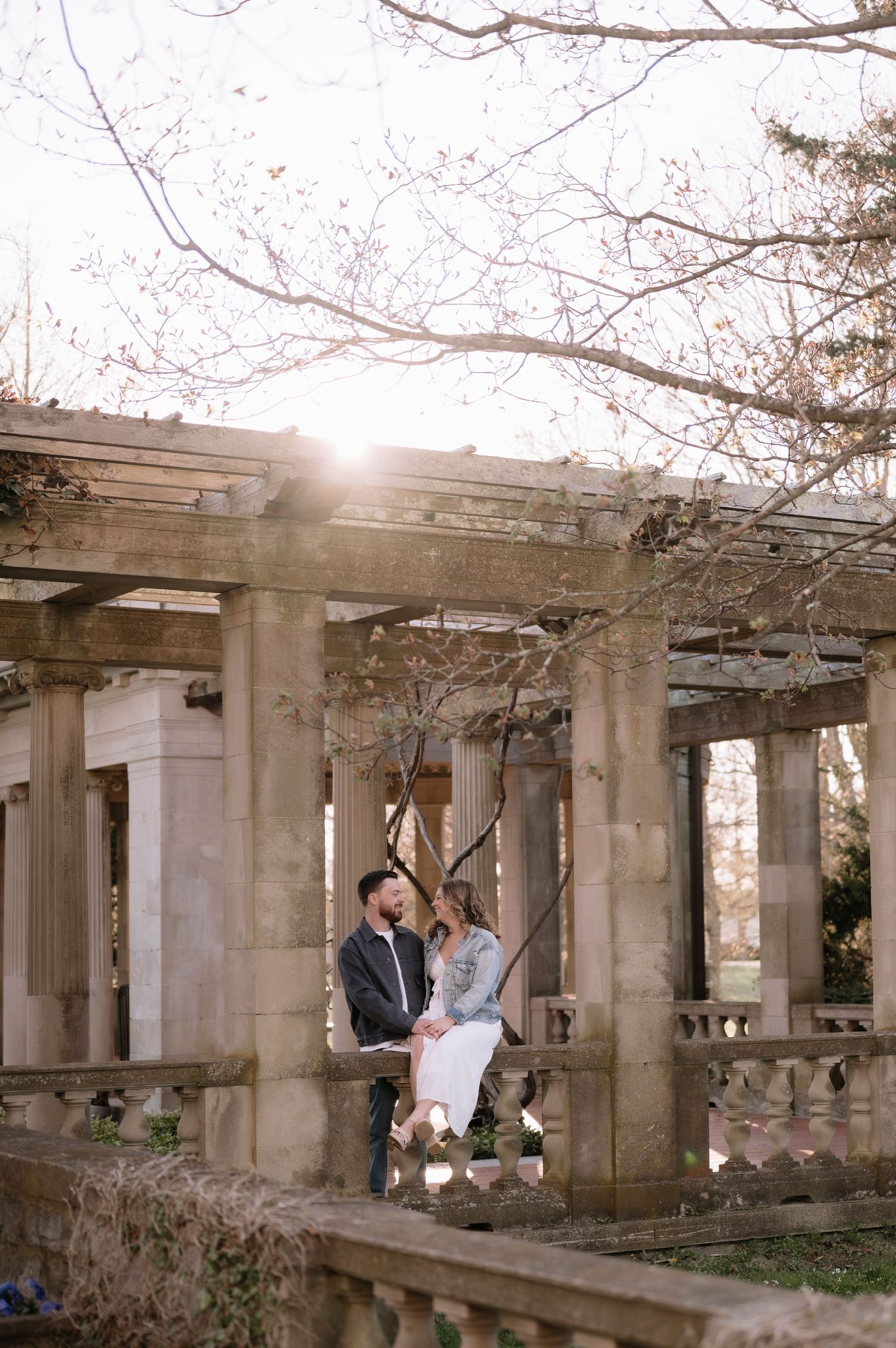 Couple sitting together among stone columns and pergola in a sunlit garden during an outdoor engagement session.