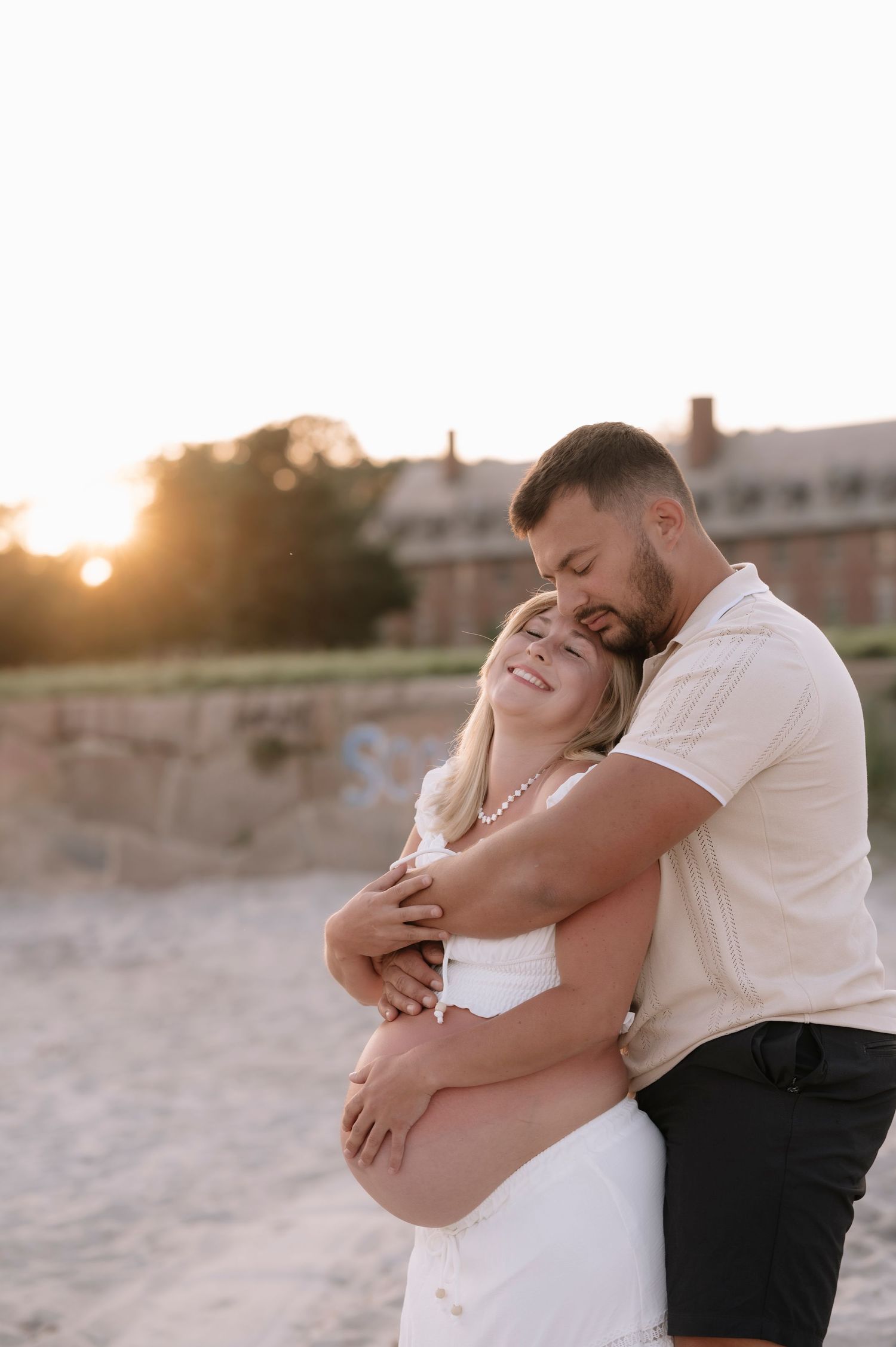 Couple embracing on sandy beach at sunset, man kissing womans head while she smiles in white dress.