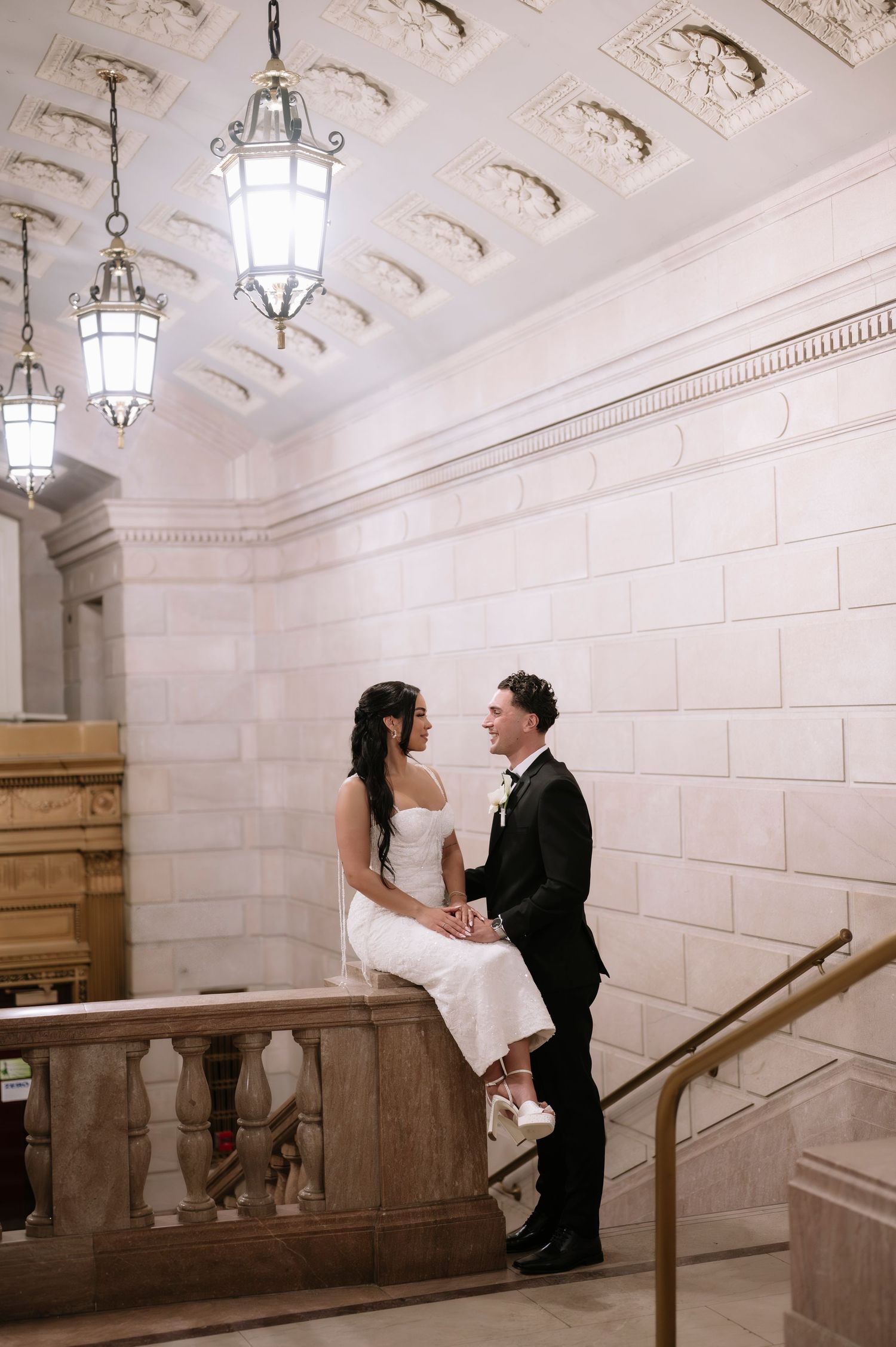 Bride sits on a marble balustrade as groom stands close, sharing an intimate moment beneath ornate ceiling lanterns.
