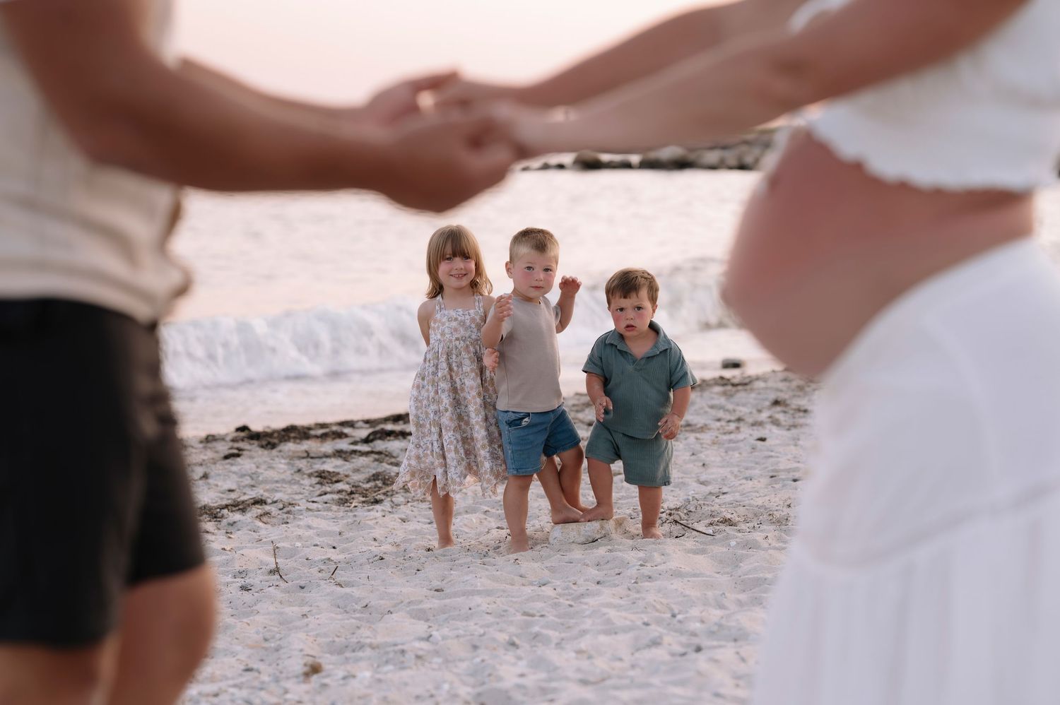 Three young children stand on a sandy beach while their parents hold hands around them at sunset.