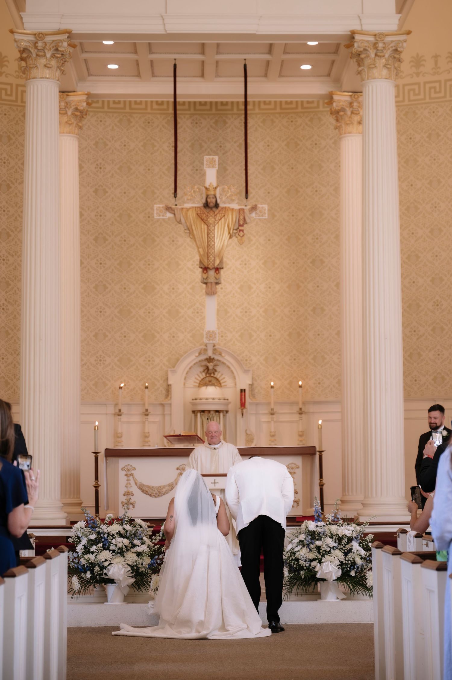 Bride and groom at church altar during wedding ceremony with ornate decor and crucifix backdrop.