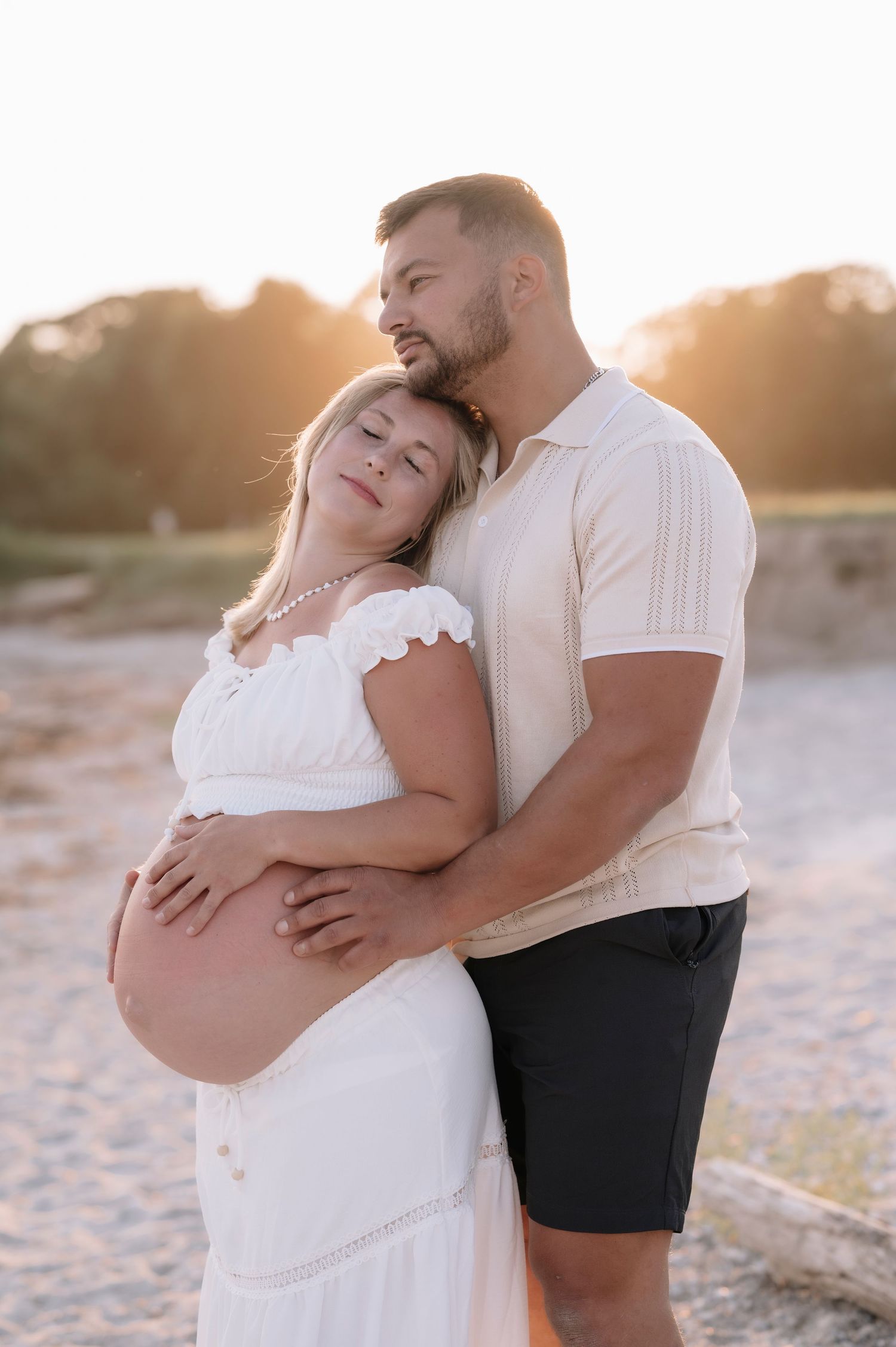 A couple shares a tender maternity photo at sunset on a beach, the woman in a white dress cradling her baby bump.