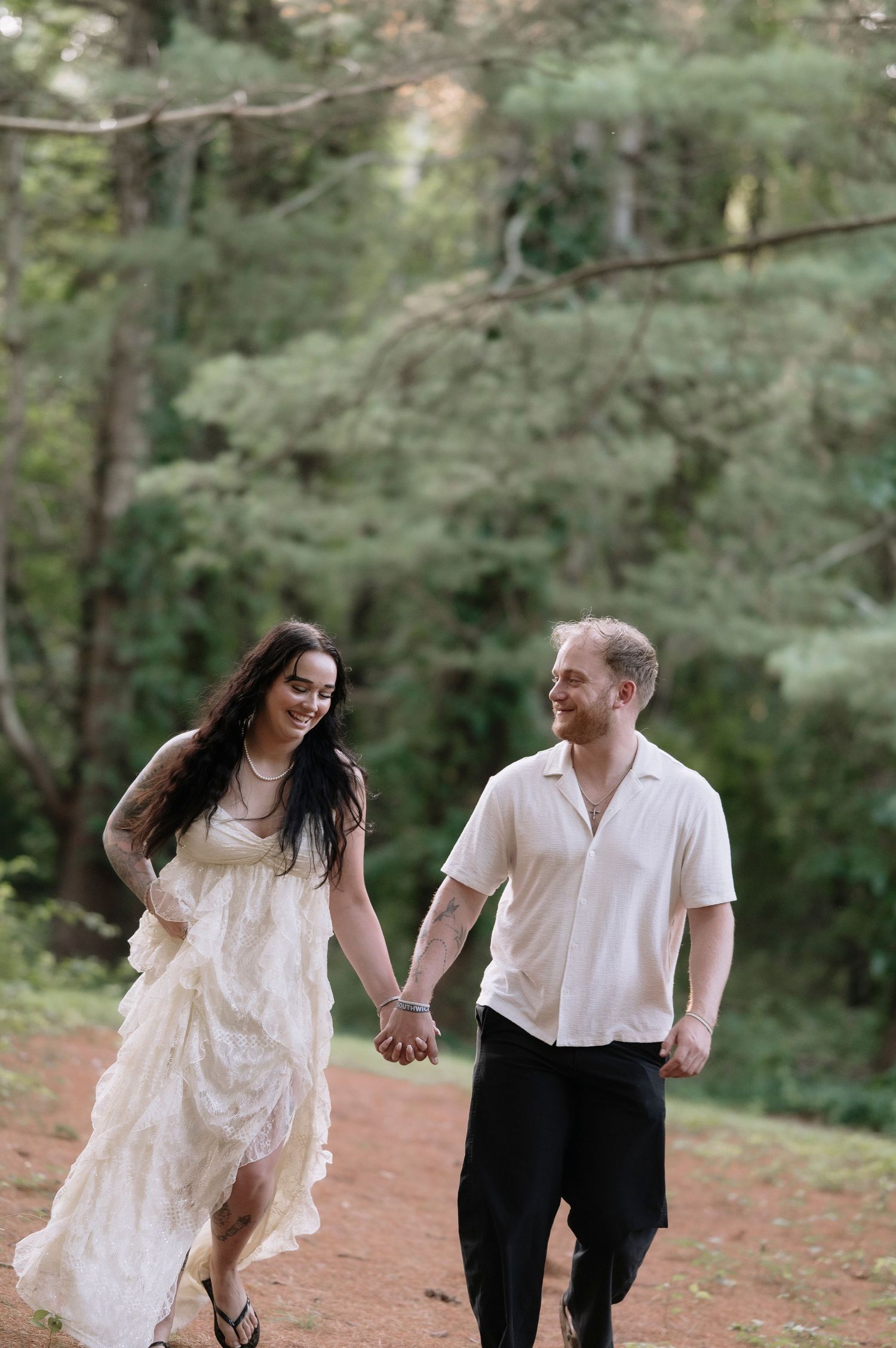 A smiling couple holds hands while walking on a dirt path surrounded by lush green trees, woman in flowing white dress.