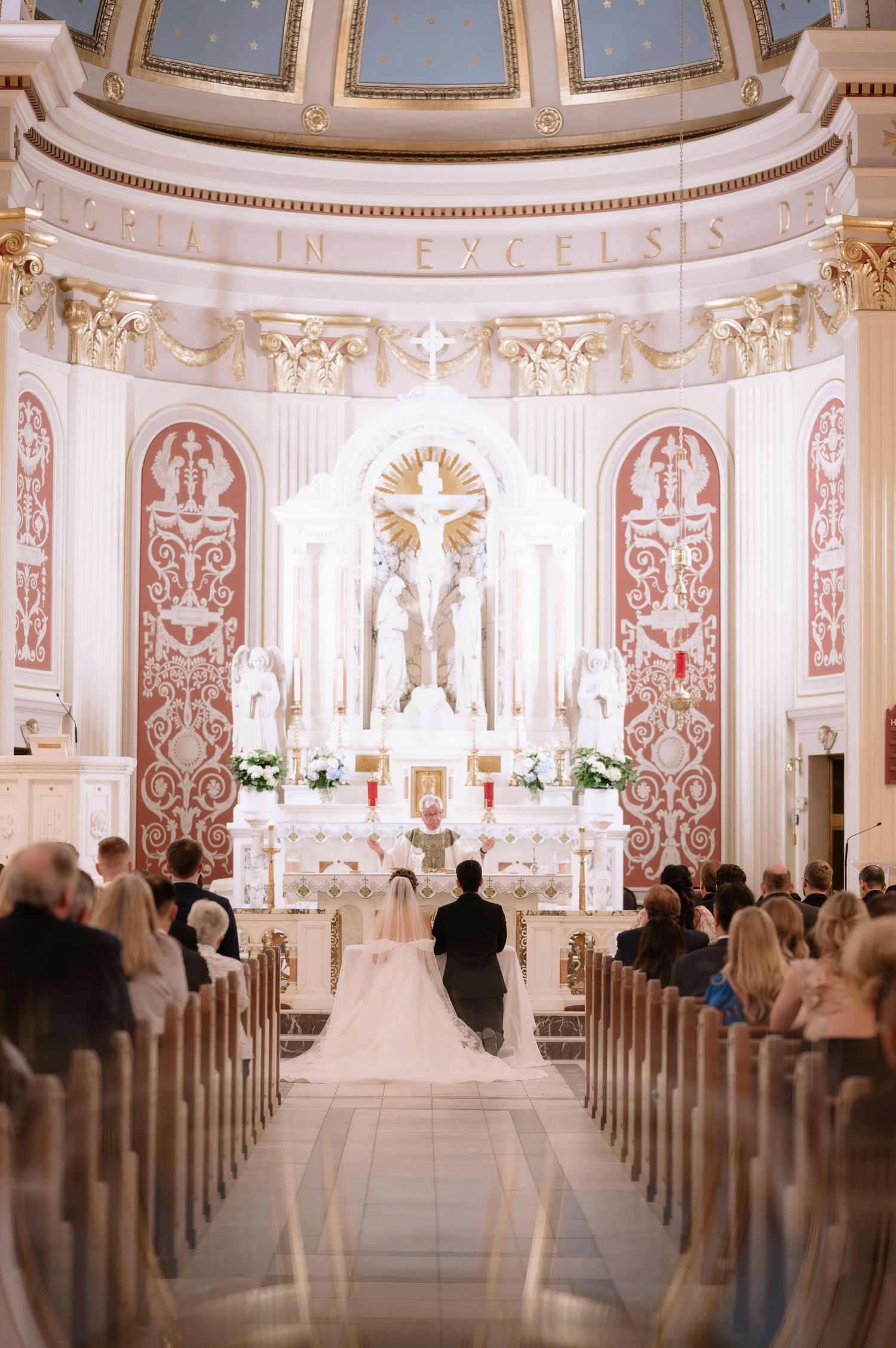 Wedding ceremony taking place in an ornate Catholic church with white marble altar and pink decorative patterns on walls.