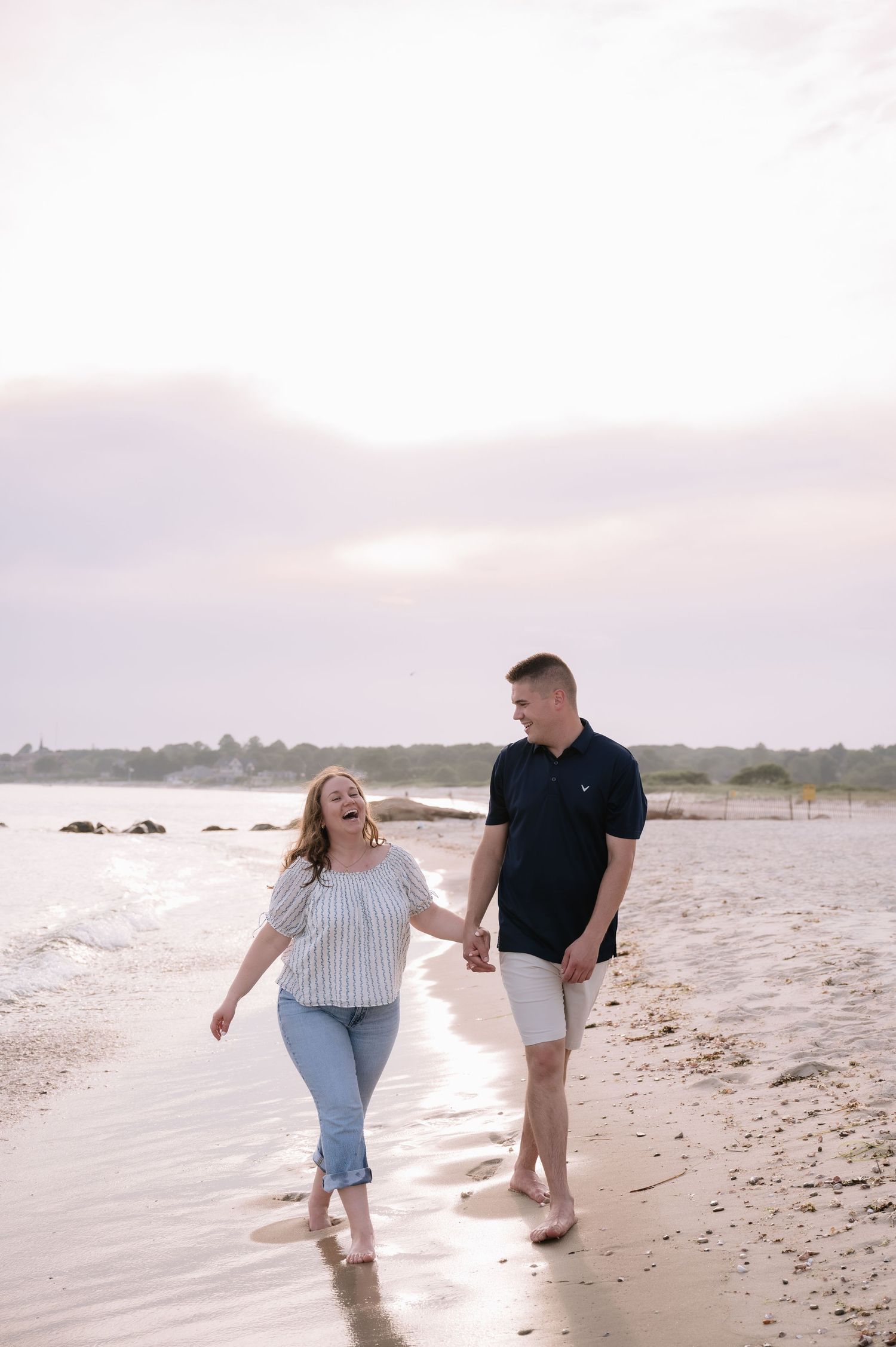 A couple walks hand in hand along a sandy beach at sunset, laughing and enjoying the ocean waves.