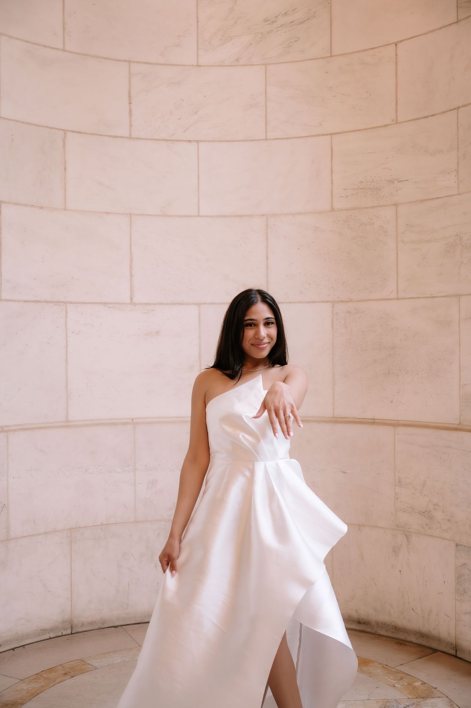 An elegant white strapless wedding gown with asymmetrical draping is showcased against a curved marble wall.