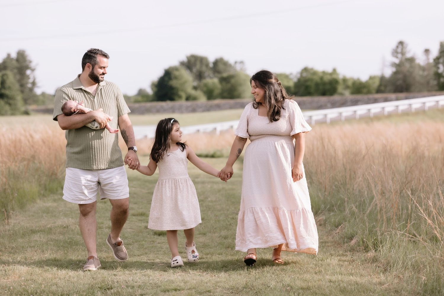 Pregnant woman and man with young girl and newborn baby walking hand in hand in a sunny field.