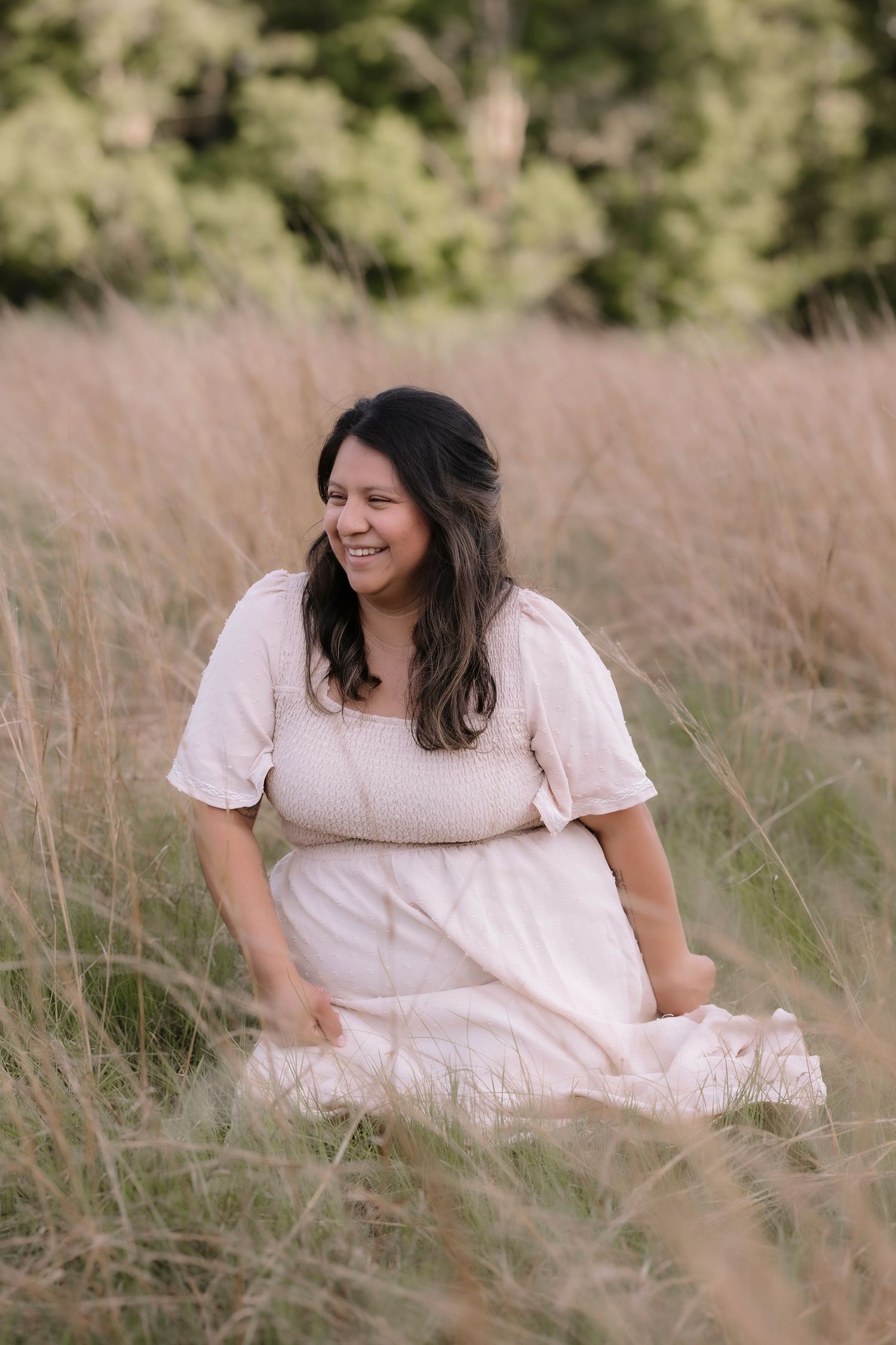 A smiling woman in a white dress sits in a golden grass field with green trees in the background.